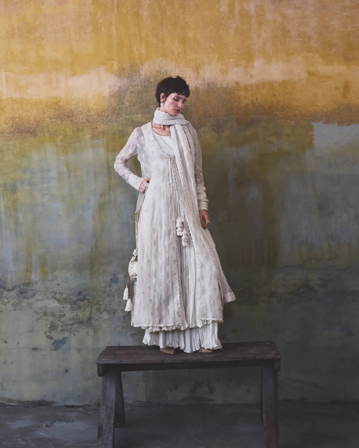 Woman in a white dress standing on a wooden bench against a textured wall.