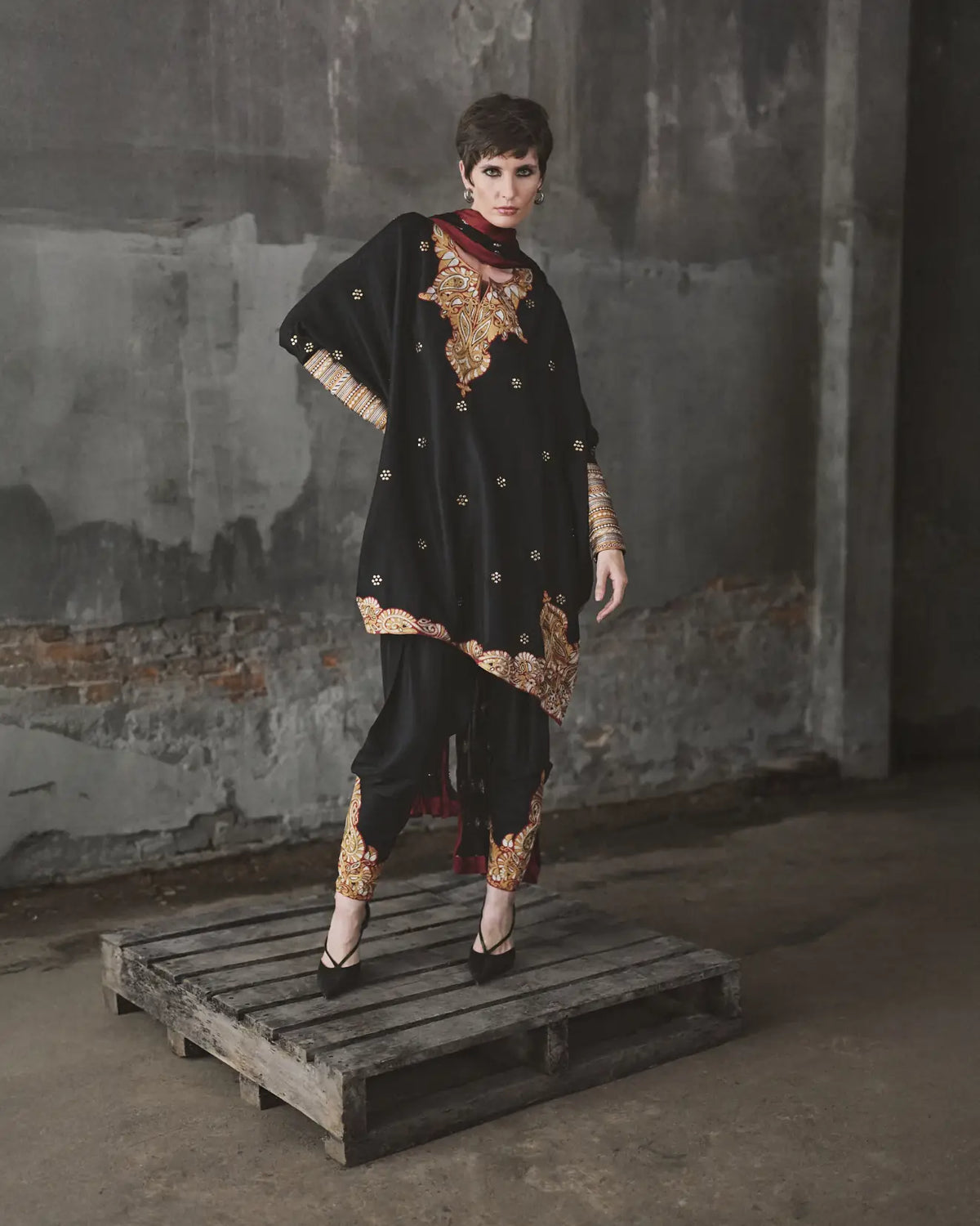 Model wearing a black and gold traditional outfit on a wooden pallet against a textured wall.