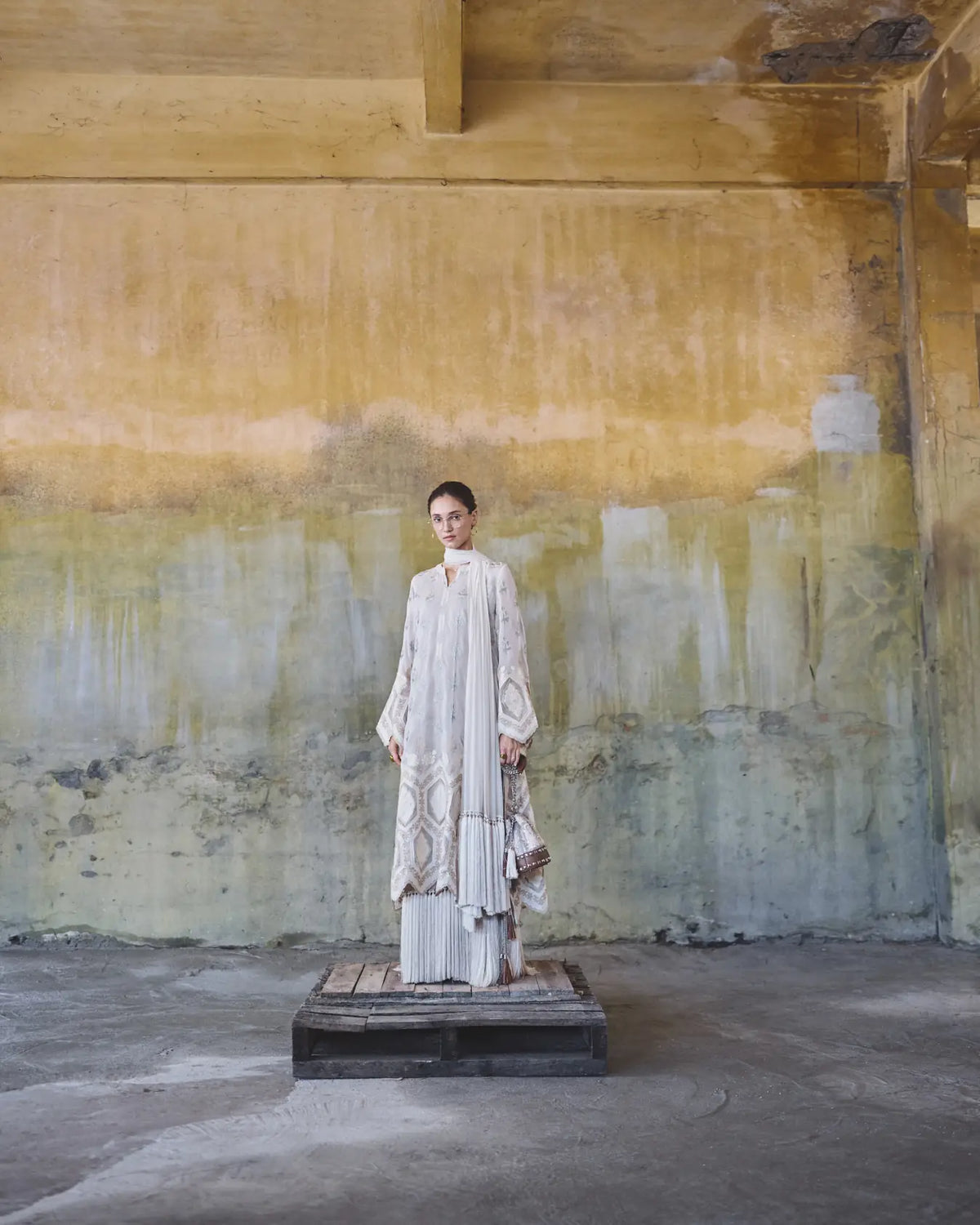 Woman in a white dress standing on a platform against a textured wall.