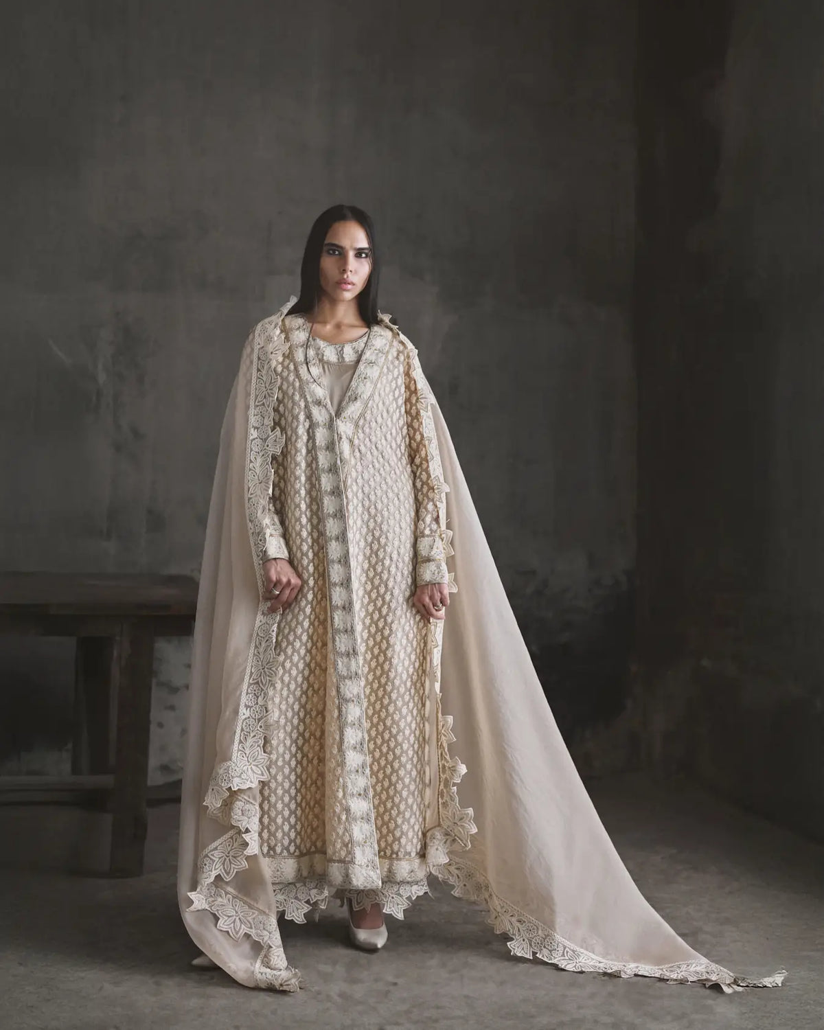Woman wearing a long, light-colored dress with a cape against a dark background