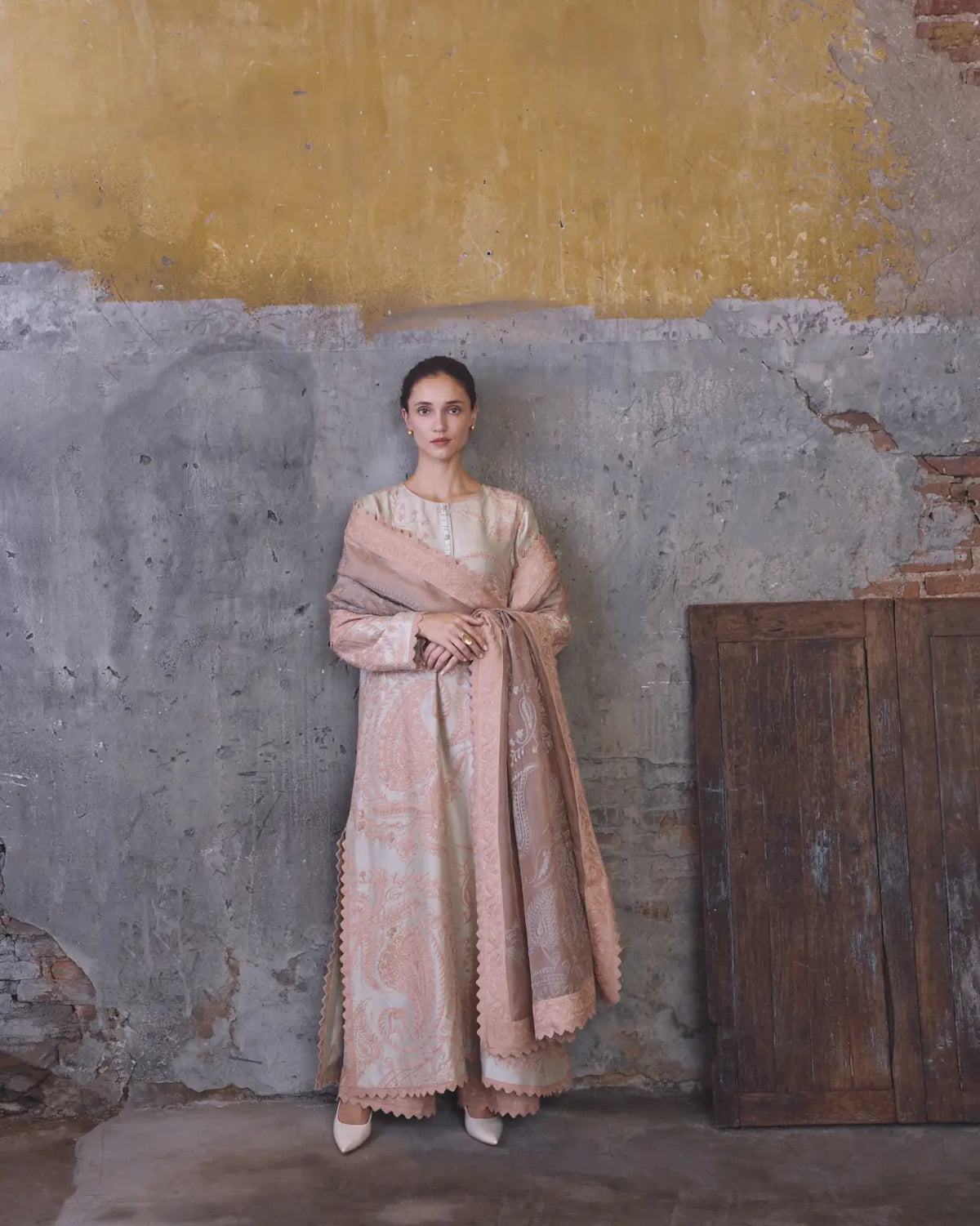 Woman in a long, patterned dress standing against a textured wall with a wooden cabinet.