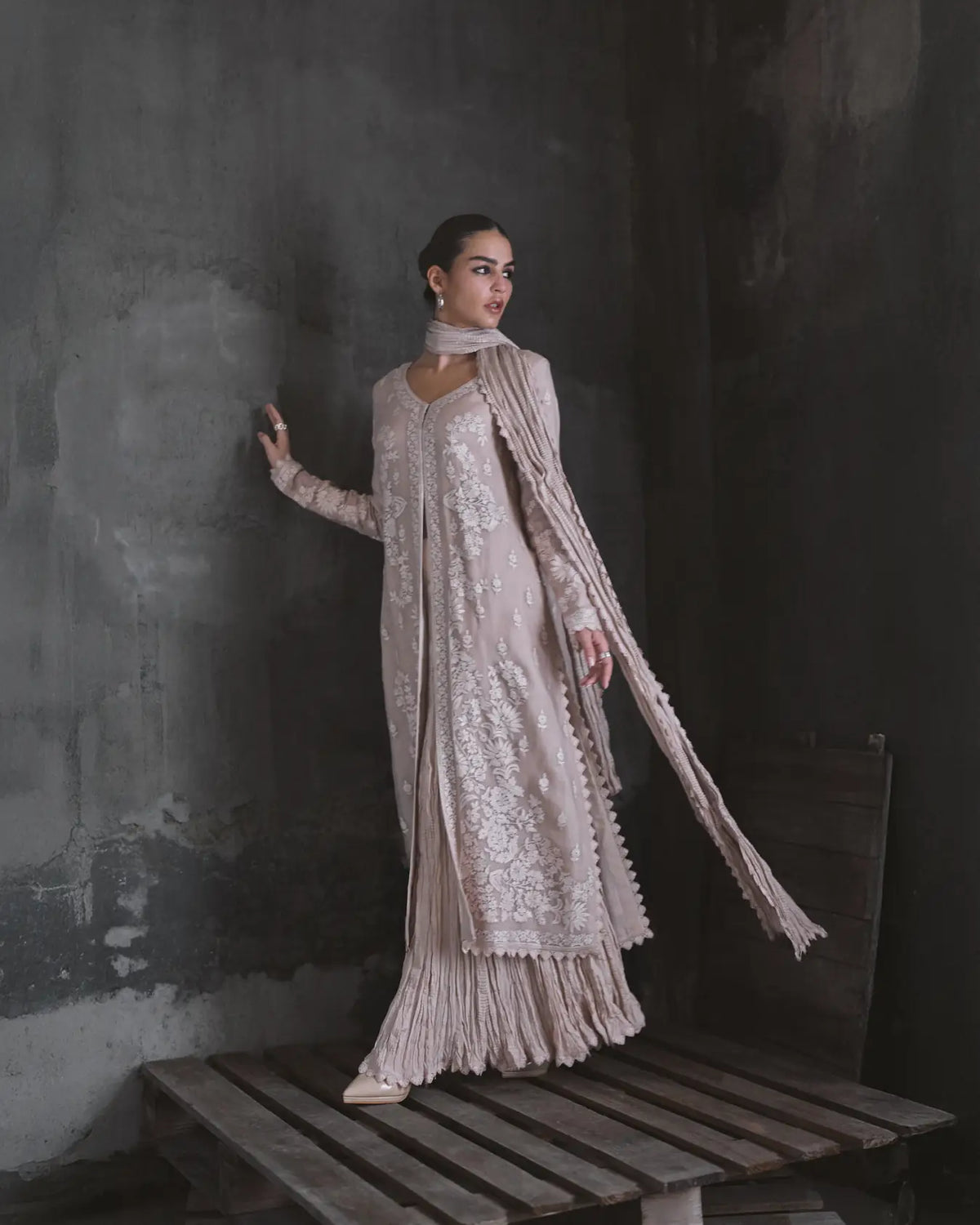 Woman in a light pink embroidered outfit standing against a dark textured wall.