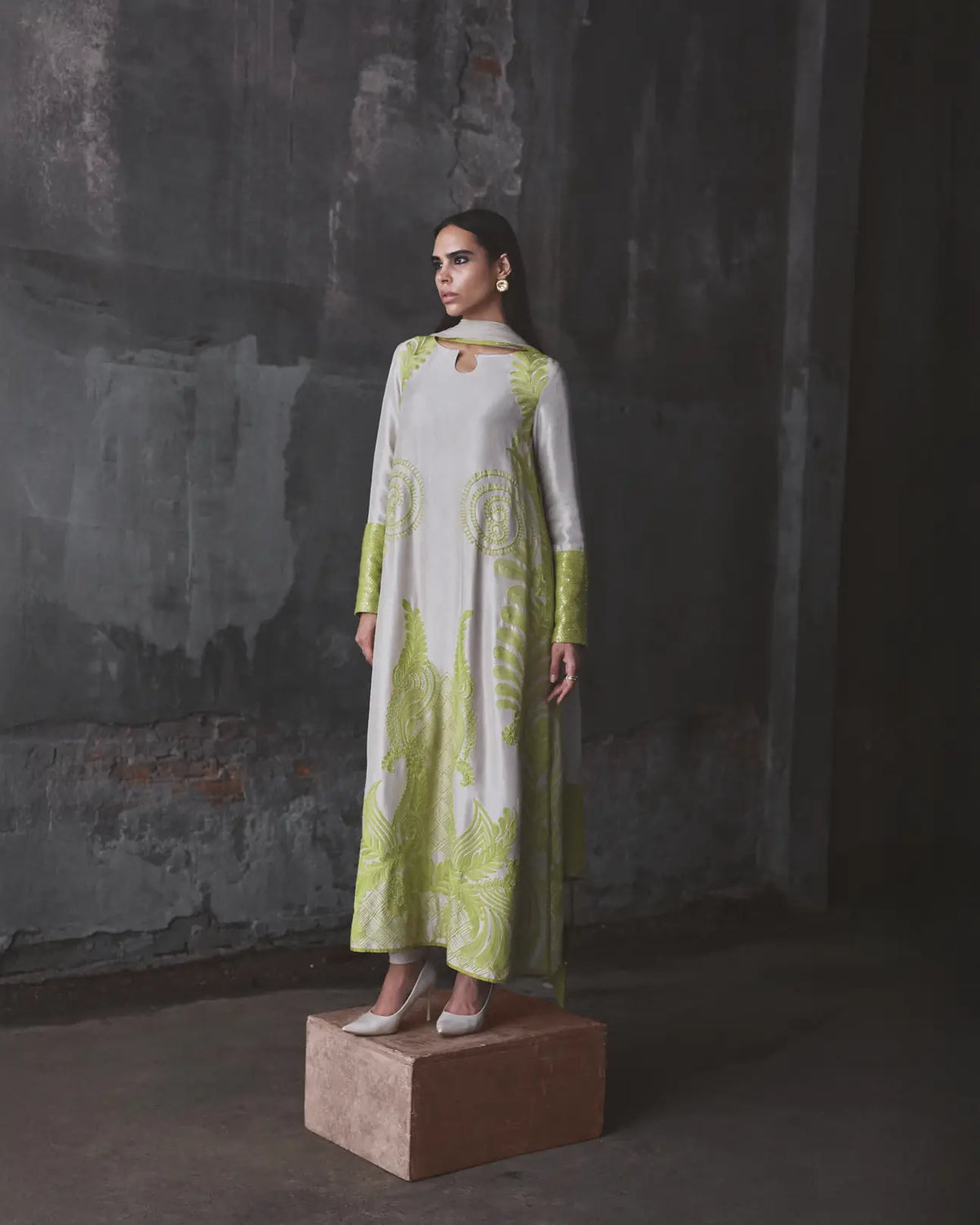 Woman wearing a light green and white patterned dress standing on a platform against a dark textured wall.