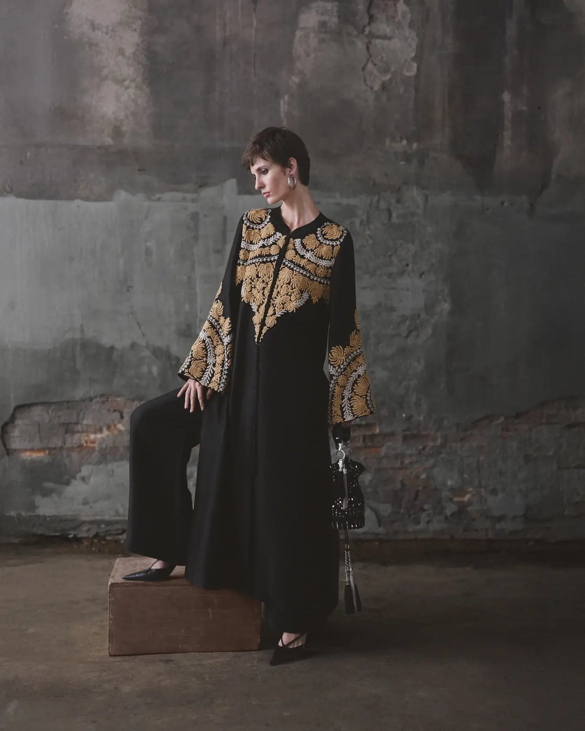 Woman in a black and gold embroidered dress sitting on a box against a textured wall.