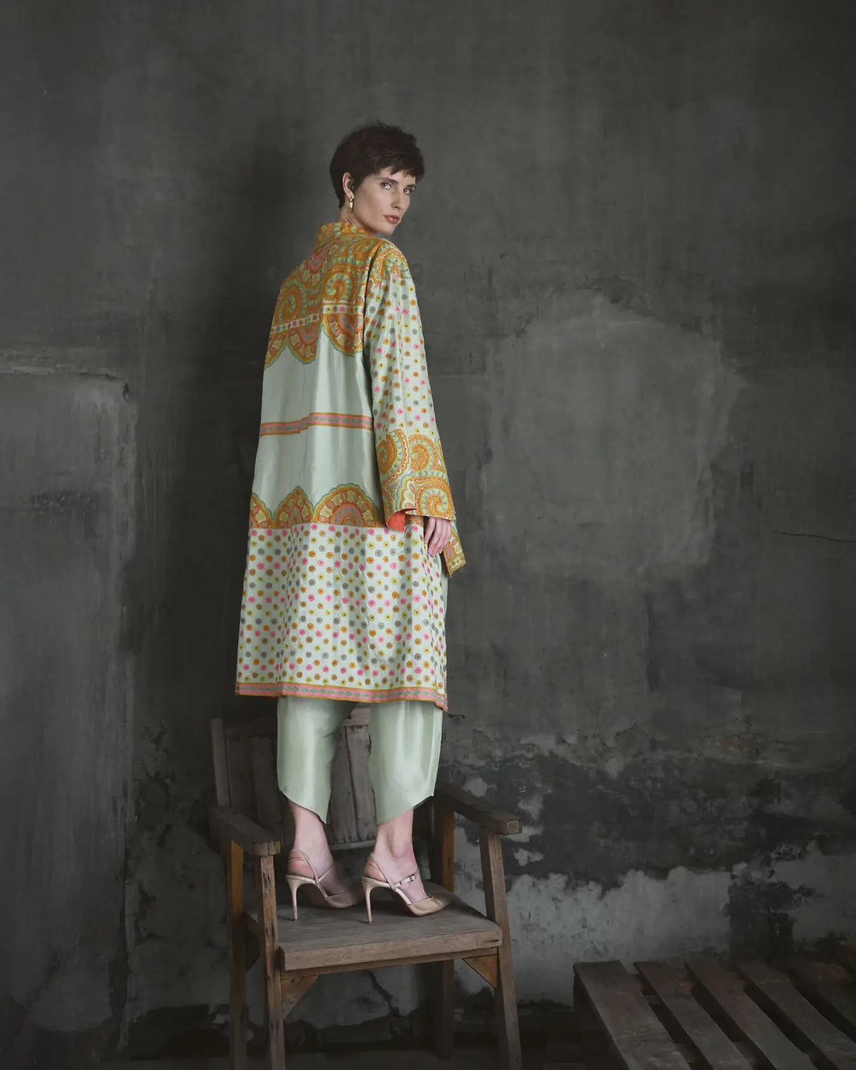 Woman in a patterned outfit standing on a chair against a textured wall.