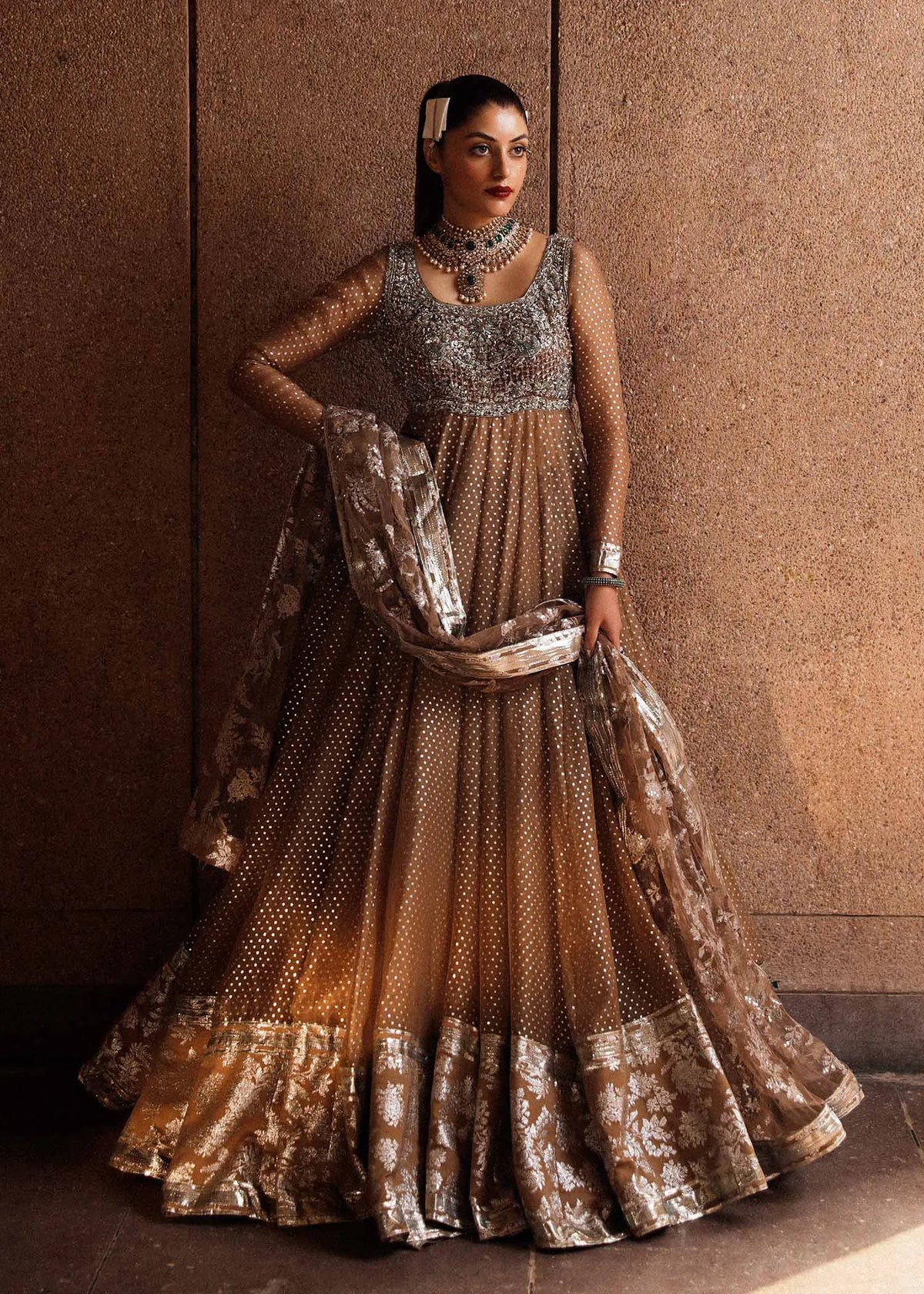 Woman in a traditional outfit with intricate design against a brown wall.