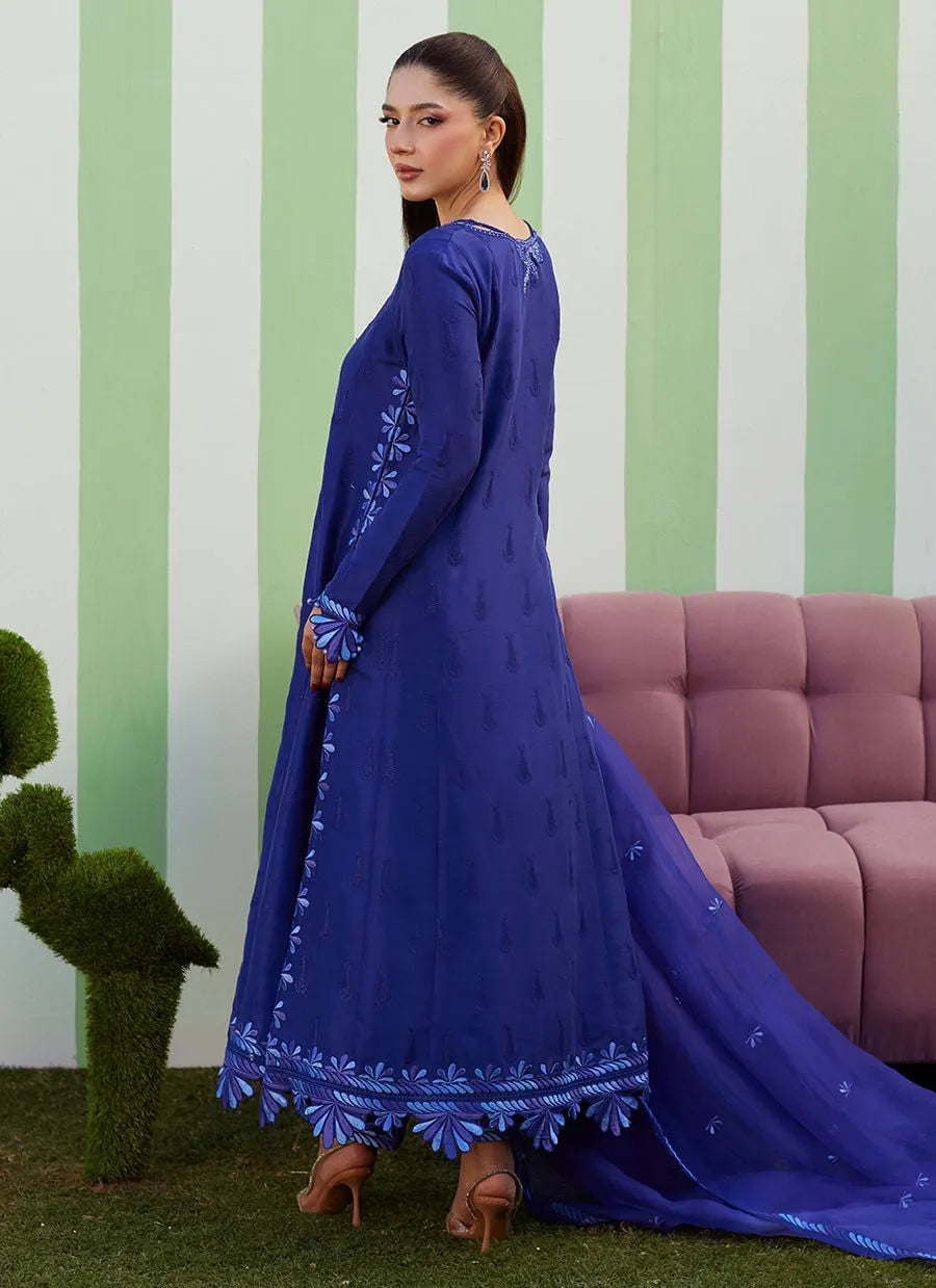 Woman in a blue embroidered traditional outfit standing in front of a striped wall with a pink sofa.