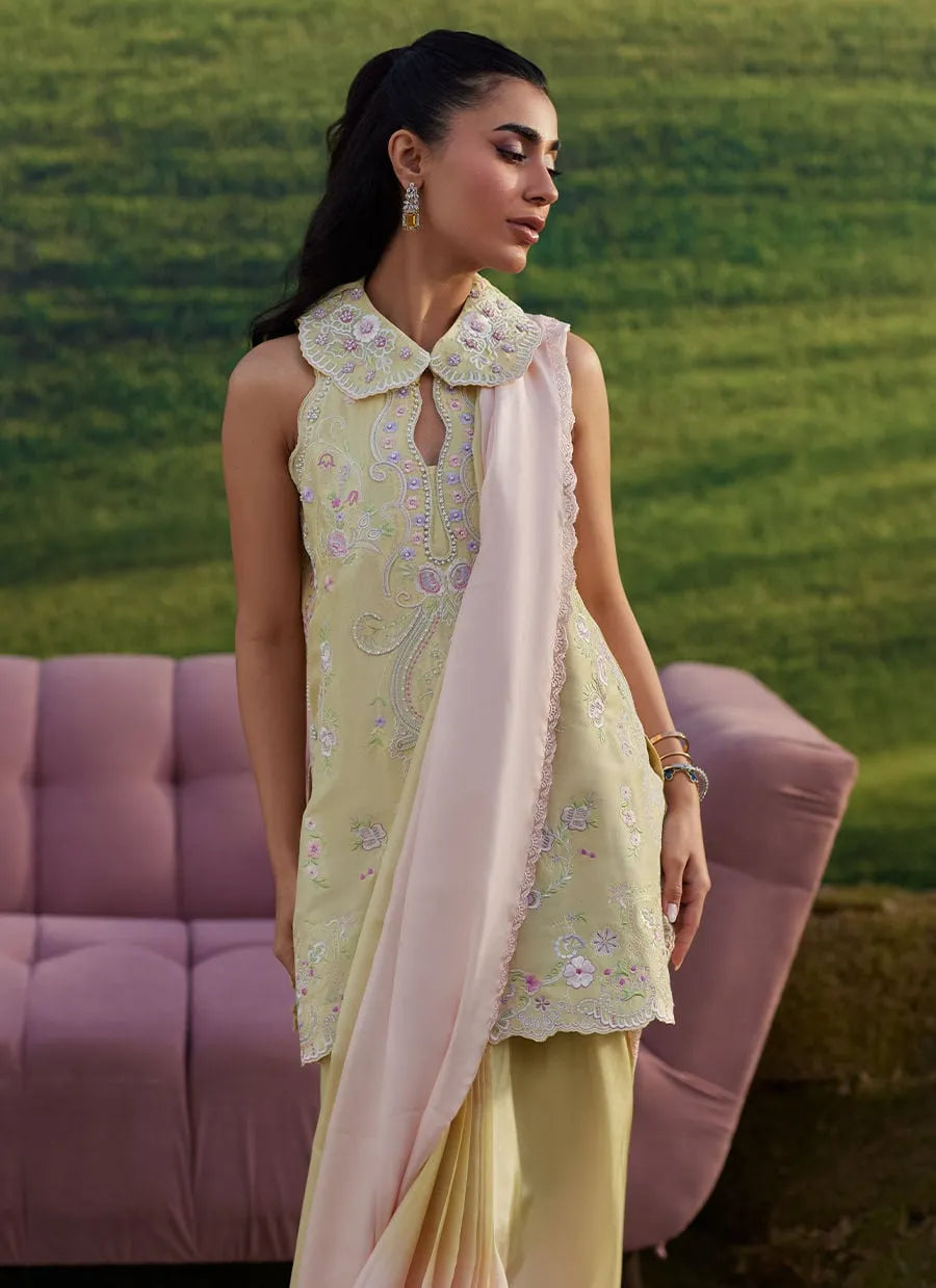 Woman in a light green embroidered outfit with a pink dupatta sitting on a pink couch against a green field.