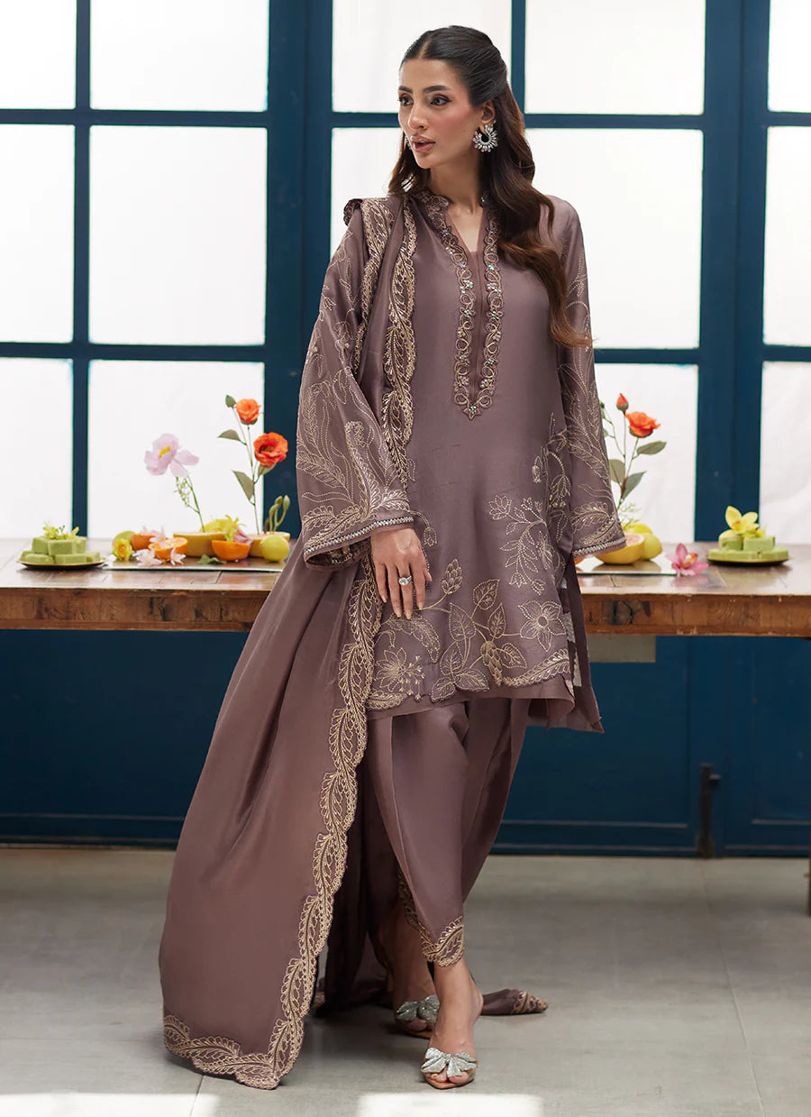 Woman in a brown embroidered outfit standing in a room with a table and flowers.