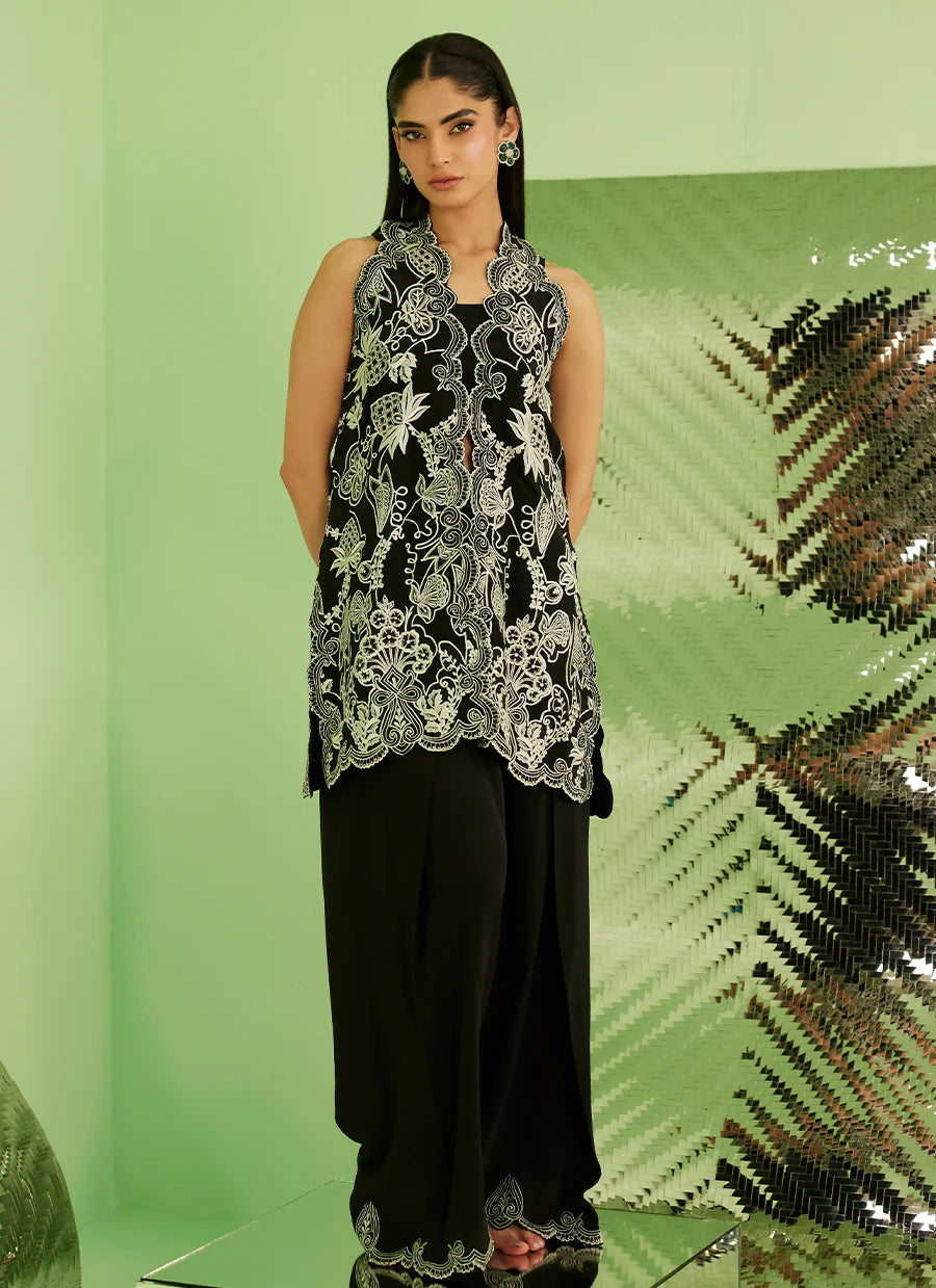 Woman wearing a black sleeveless top with white floral embroidery and black pants against a green background.