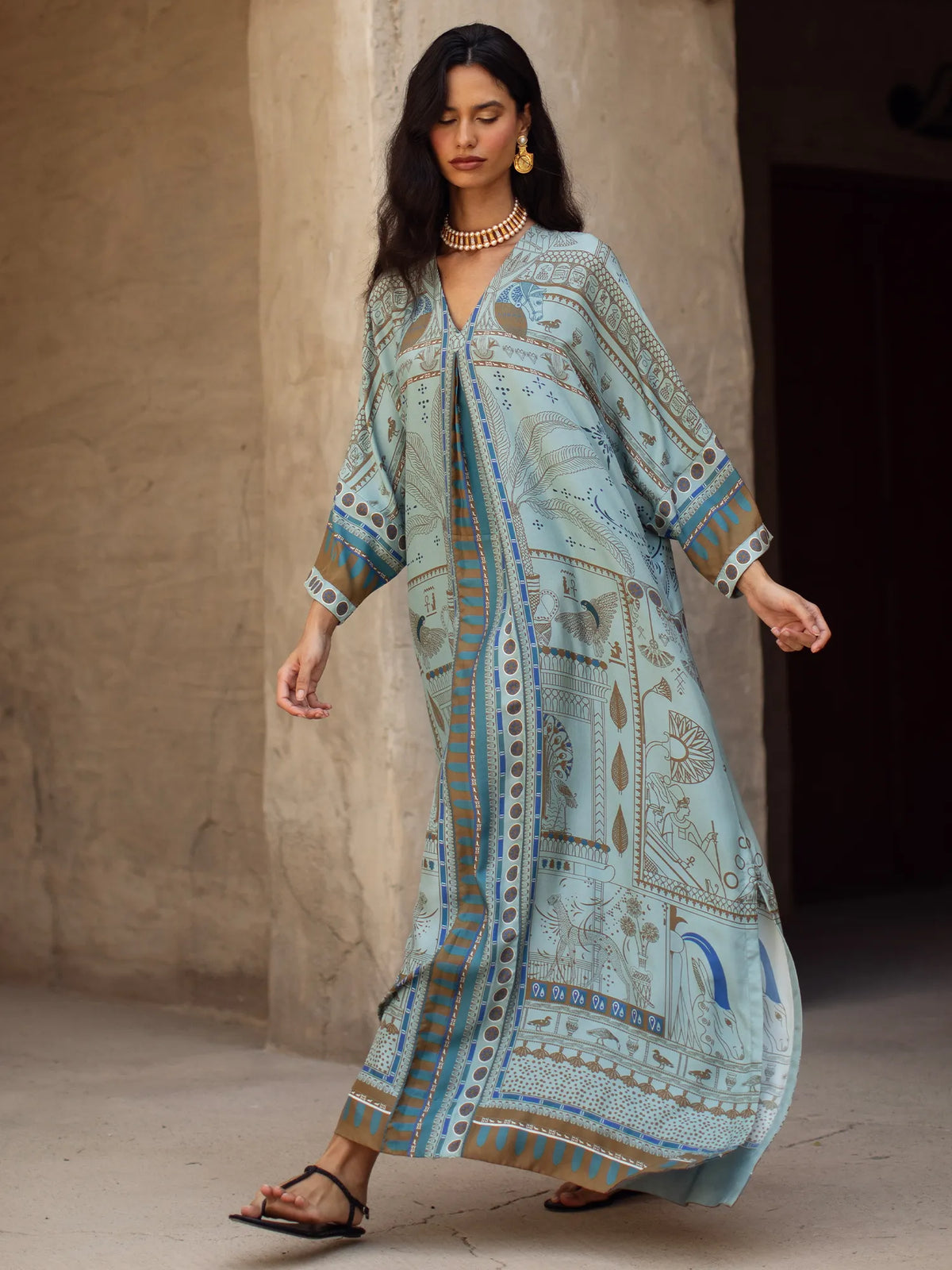 Woman wearing a long, light blue patterned dress against a plain wall.