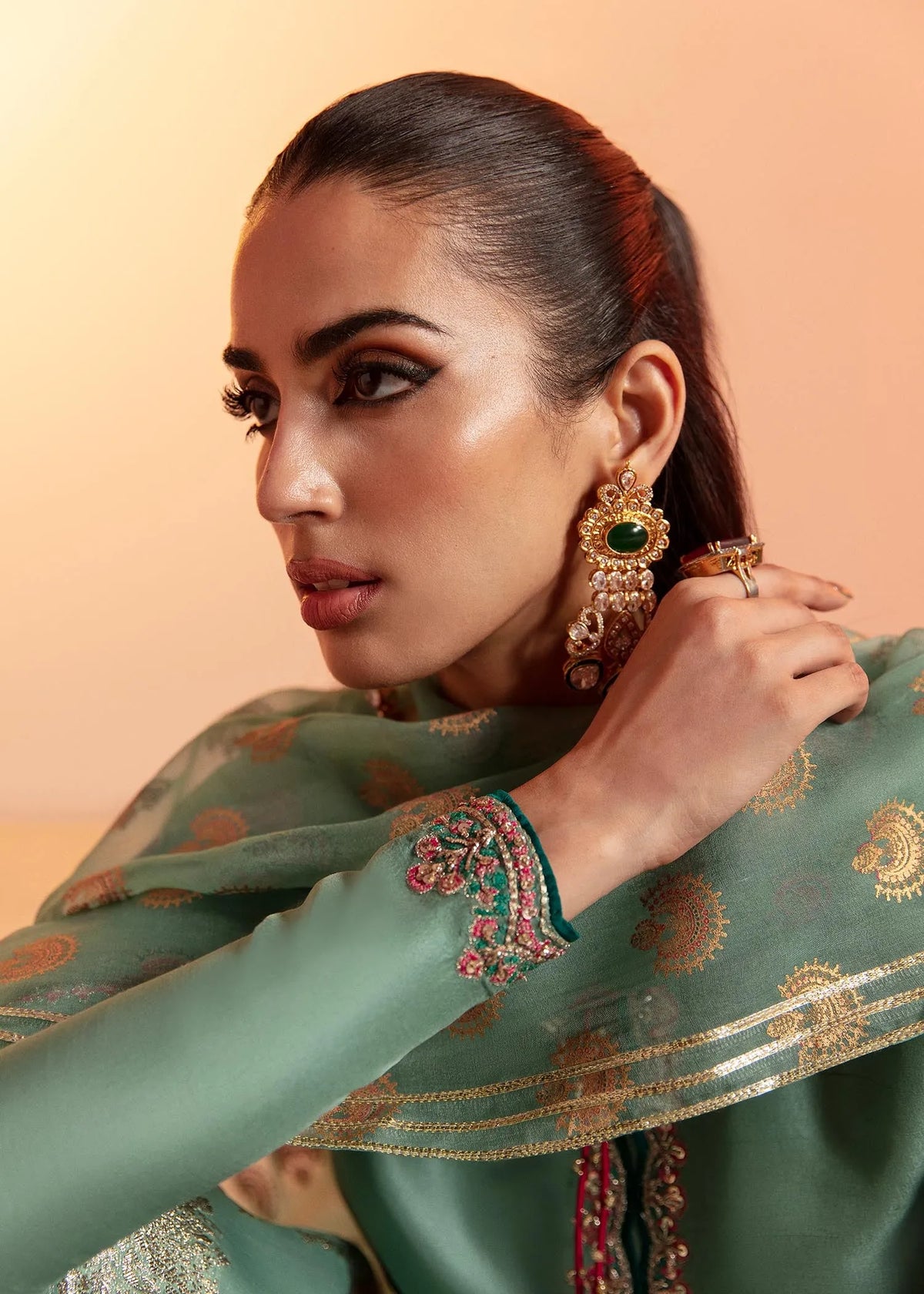 Woman wearing a green traditional outfit with gold embroidery, posing against a peach background.