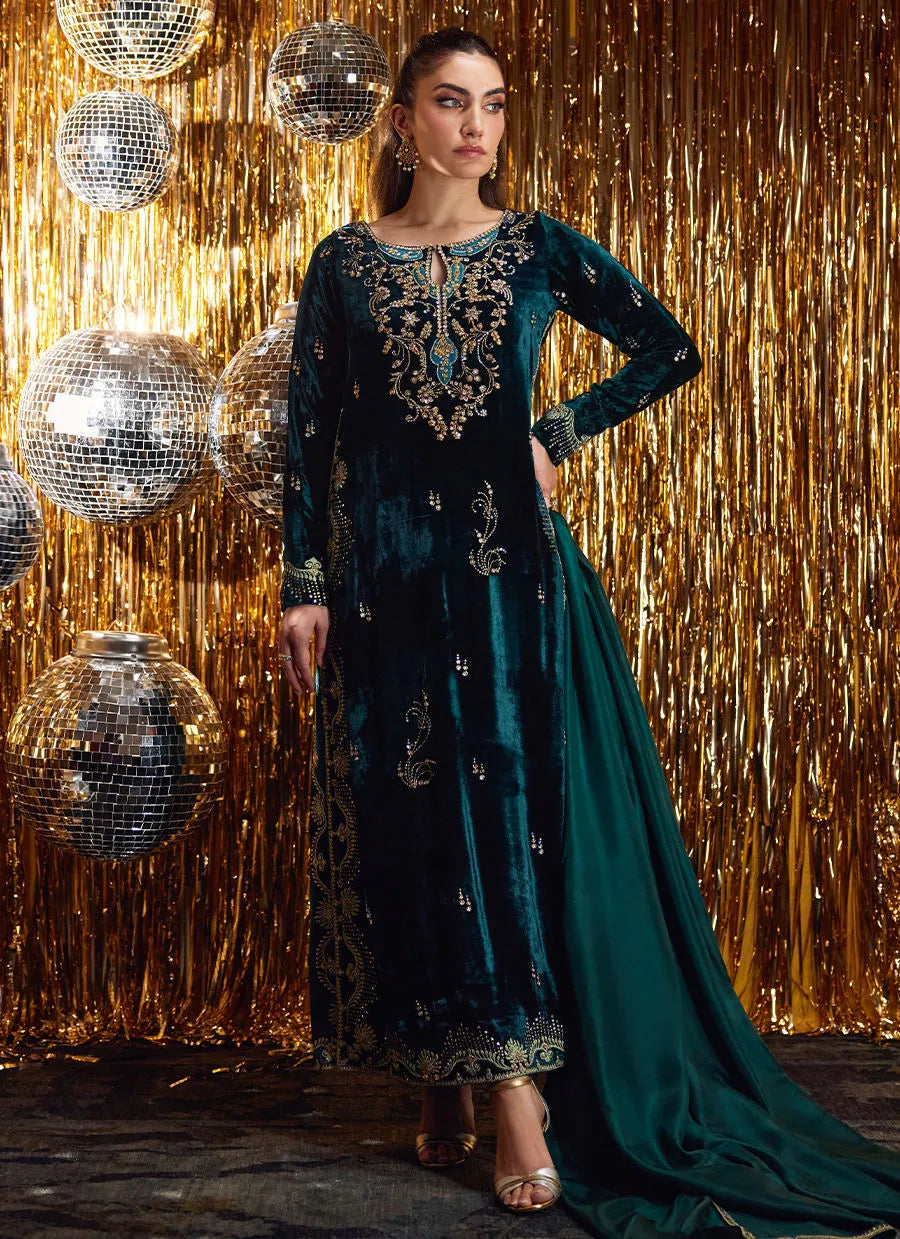 Woman in a teal embroidered dress standing against a gold glittery curtain with disco balls.