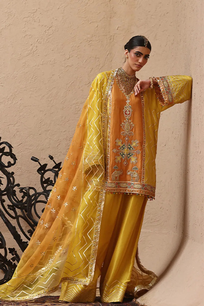 Woman wearing a traditional yellow and orange embroidered outfit against a beige wall.