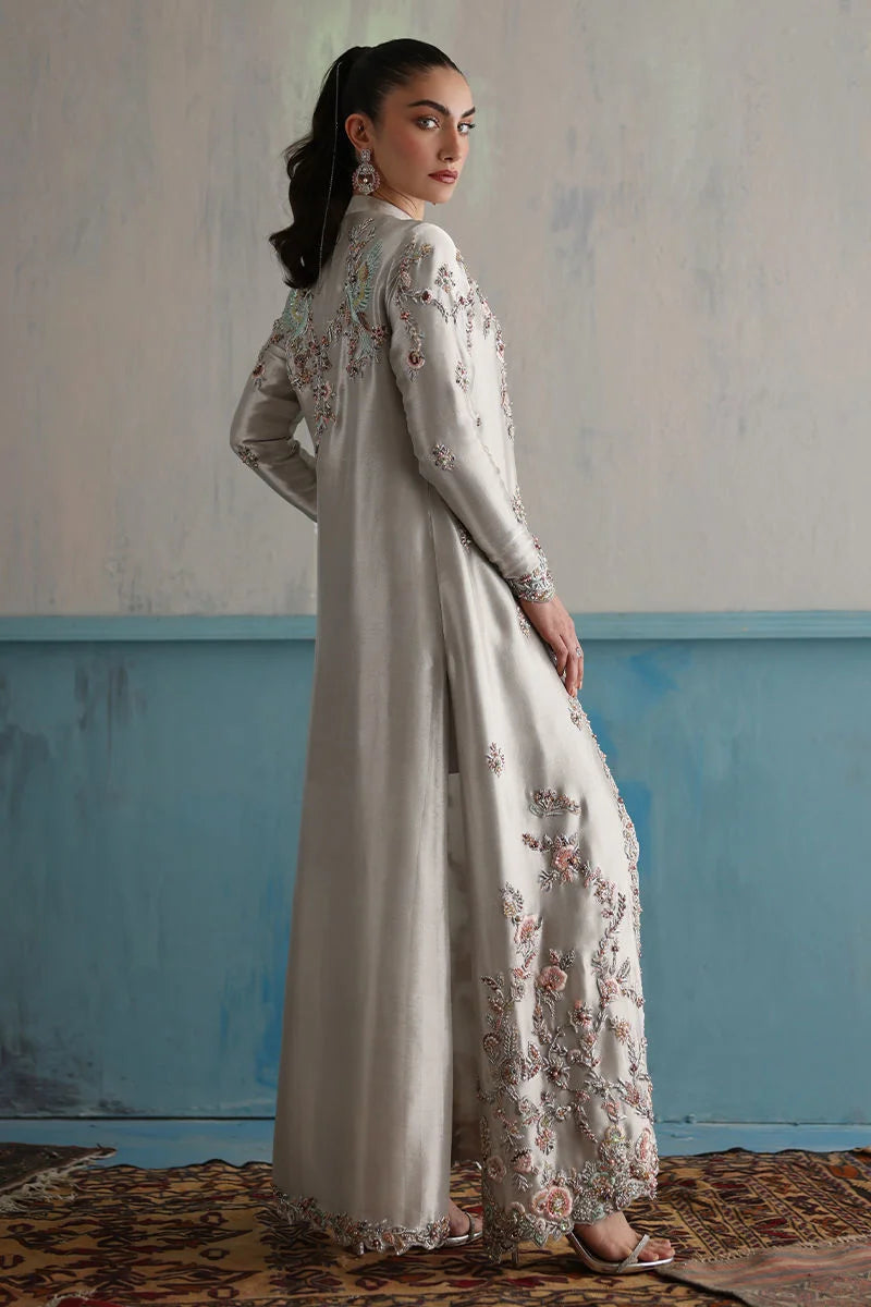 Woman wearing a long, light-colored embroidered dress against a textured wall with blue baseboard.