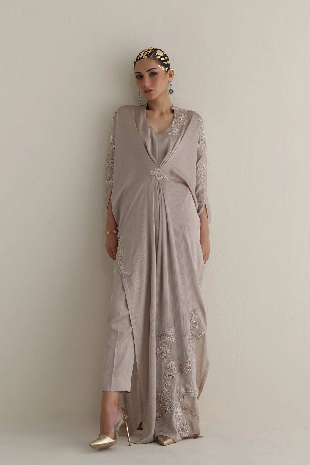 Woman wearing a long, light-colored dress with floral embroidery against a plain background