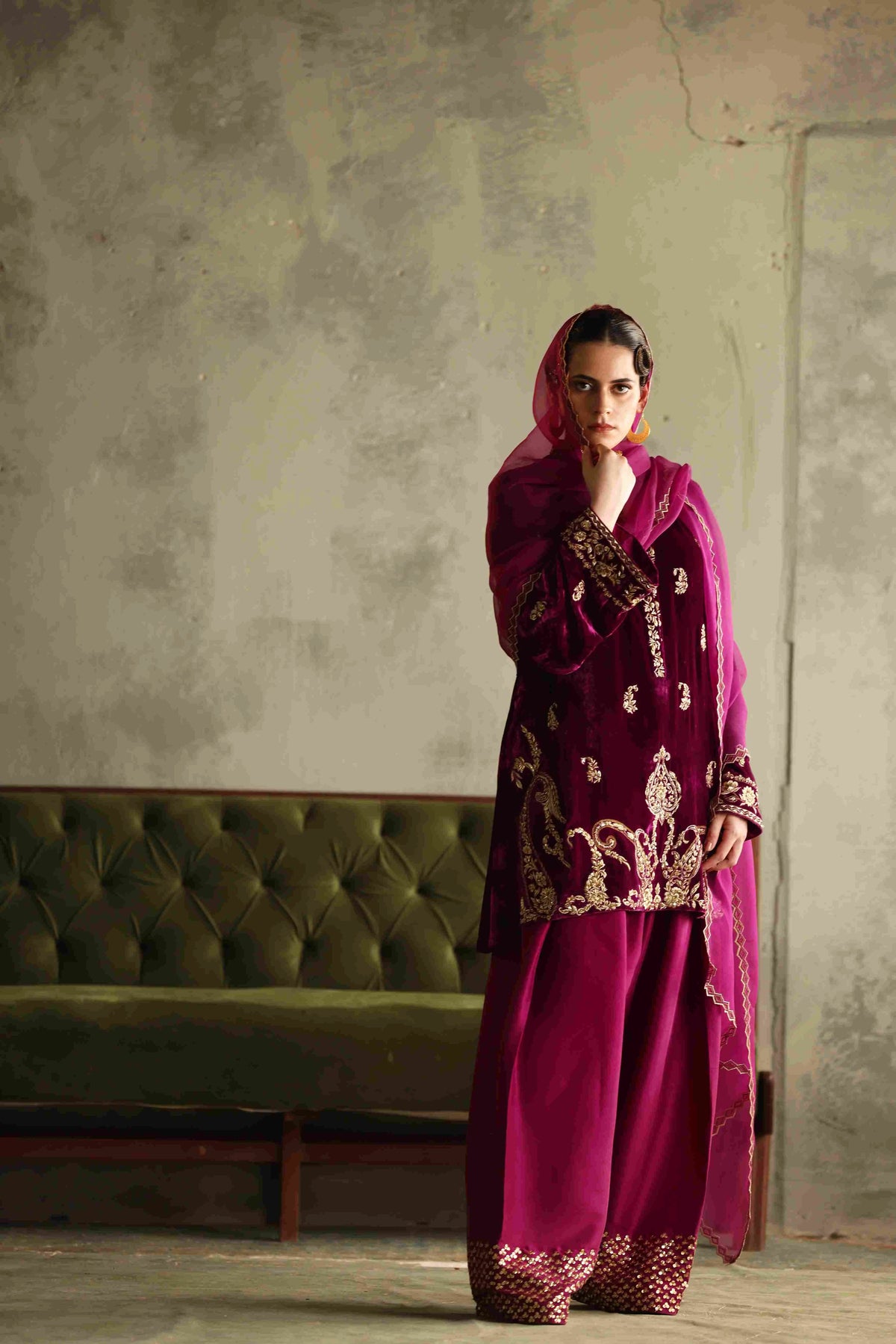 Woman in a magenta embroidered outfit standing against a textured wall with a green sofa.