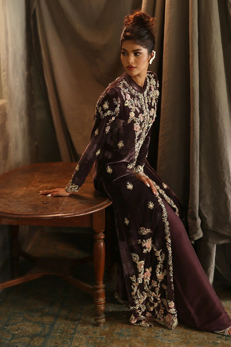Woman in a dark purple embroidered outfit sitting on a wooden stool against a draped curtain background.
