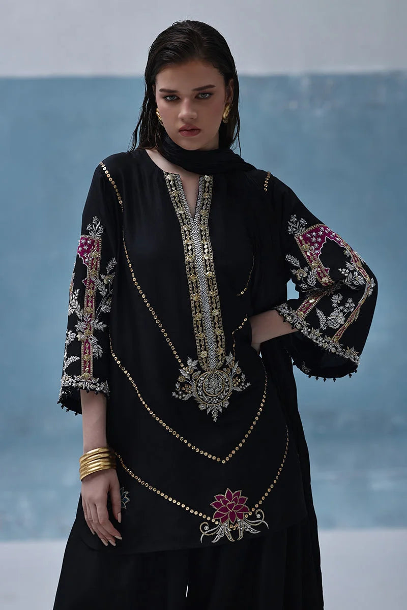 Woman wearing a black embroidered dress with floral patterns against a blue and gray background