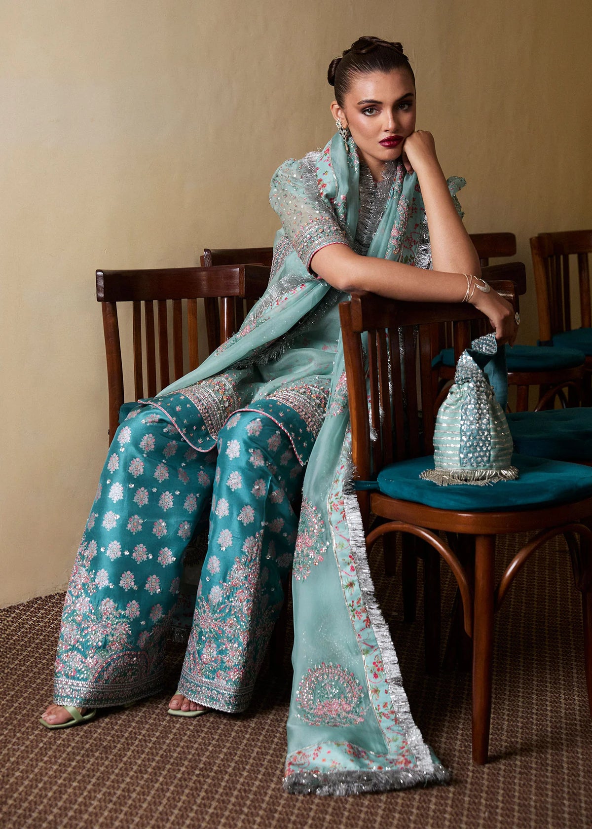 Woman in a teal and silver traditional outfit sitting on a wooden chair.