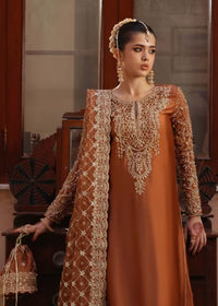 Woman in an orange traditional outfit with intricate gold embroidery, standing indoors.