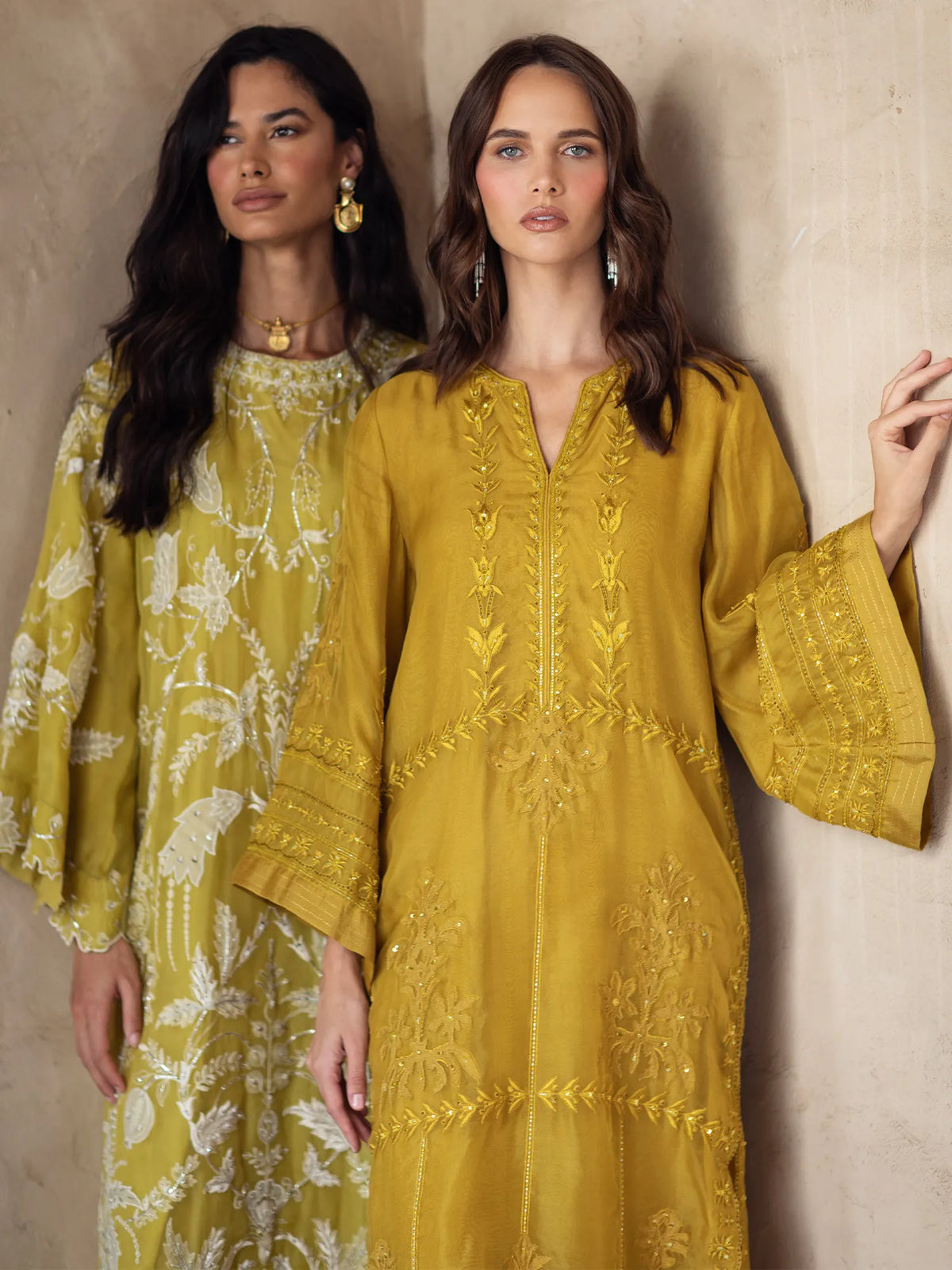 Two women wearing yellow embroidered dresses against a beige wall.
