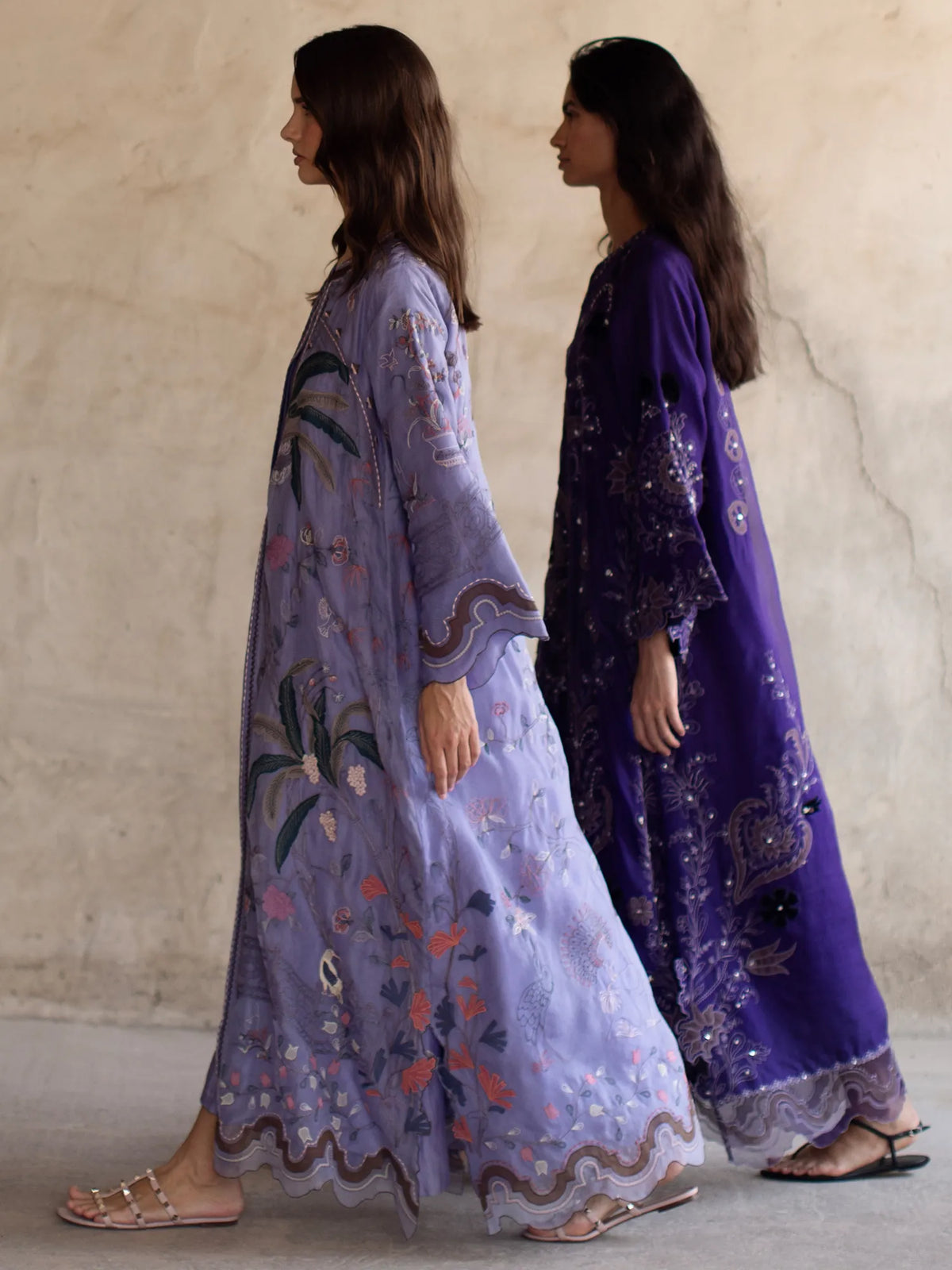 Two women wearing embroidered dresses against a neutral wall.