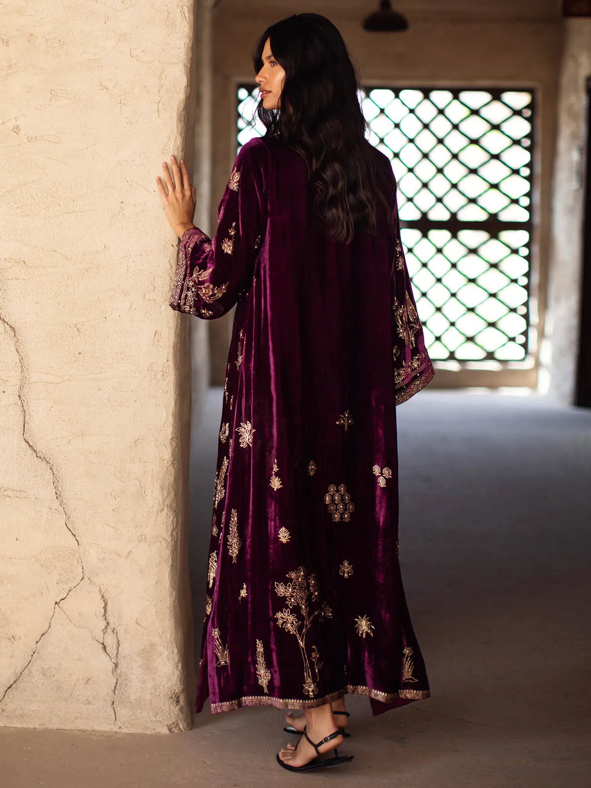 Woman in a long, dark purple dress with floral patterns standing against a textured wall.