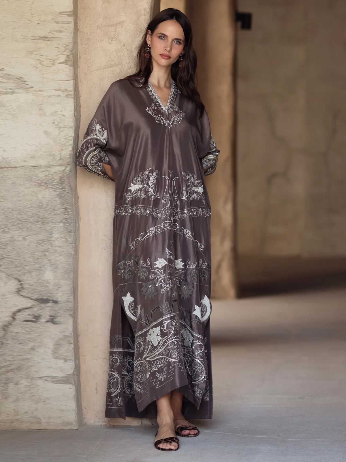 Woman wearing a long, dark brown dress with white patterns against a stone wall.