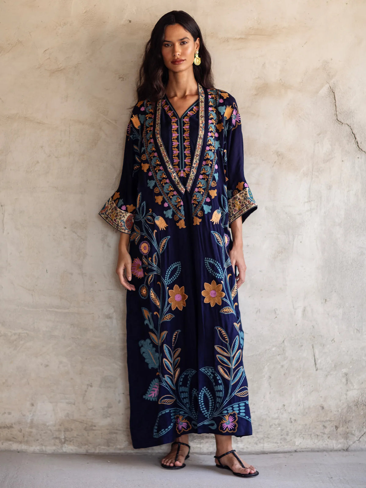 Woman wearing a long, dark blue dress with floral patterns against a light gray wall.