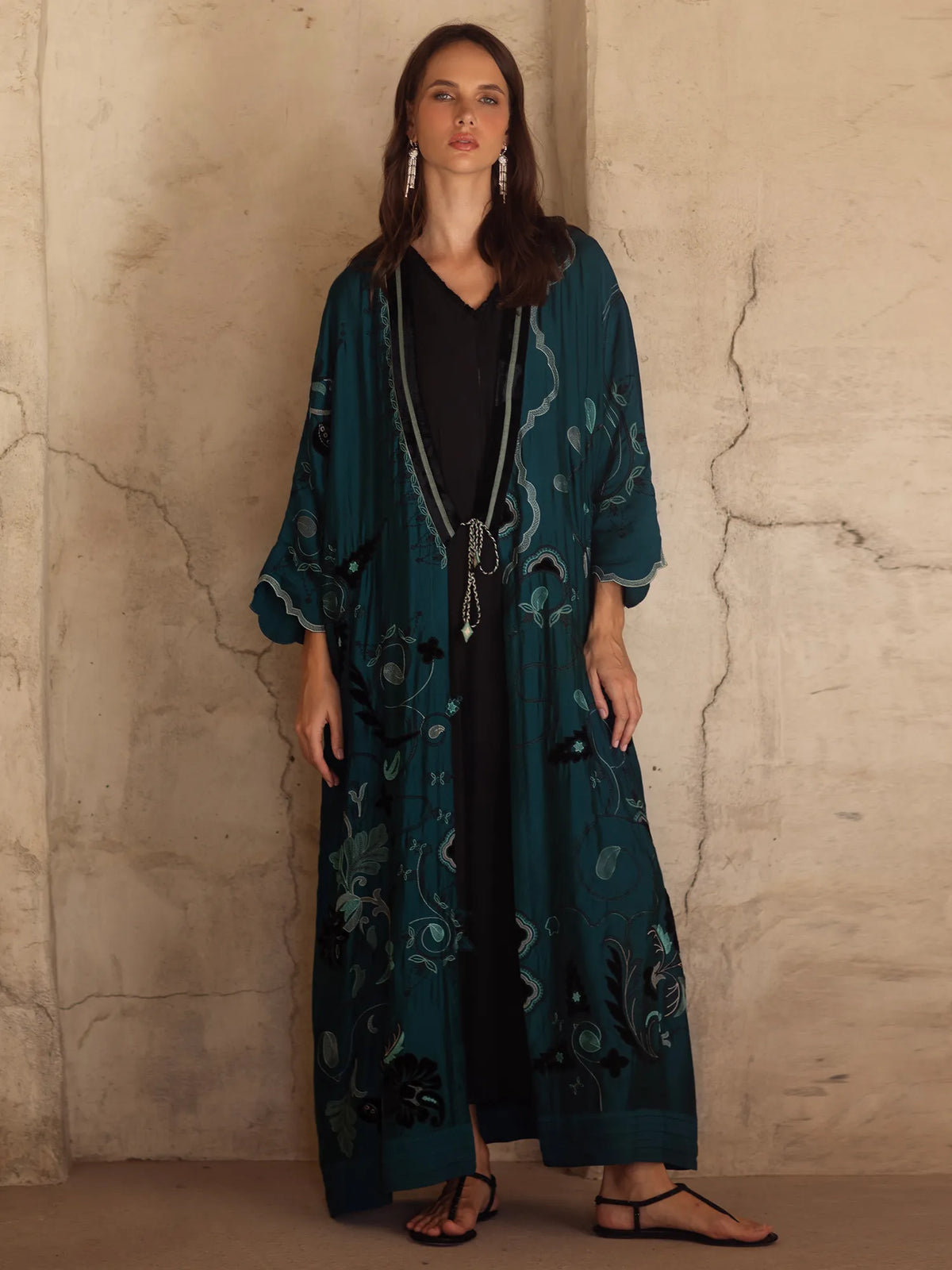 Woman wearing a long teal dress with floral patterns against a textured beige wall.