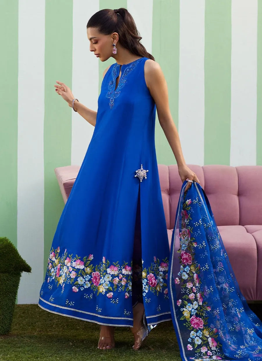 Woman in a blue dress with floral embroidery holding a matching scarf against a striped wall.