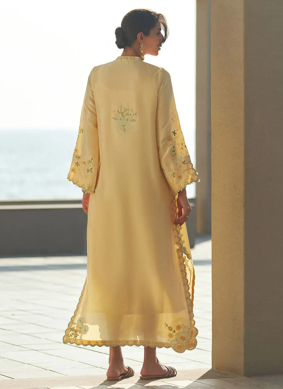 Woman in a yellow embroidered dress standing by a window with a view of water.