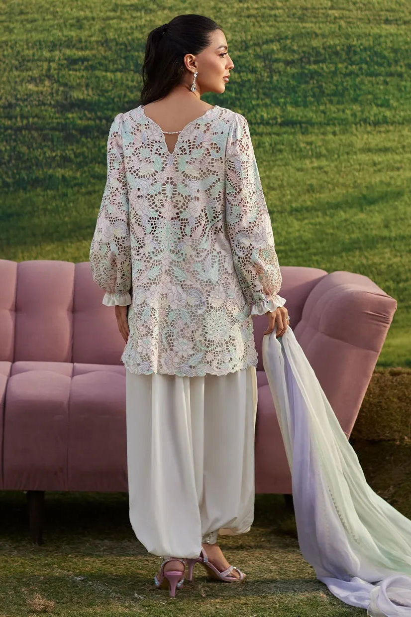 Woman in a floral top and white pants holding a dupatta, standing in front of a pink sofa with a green grass background.
