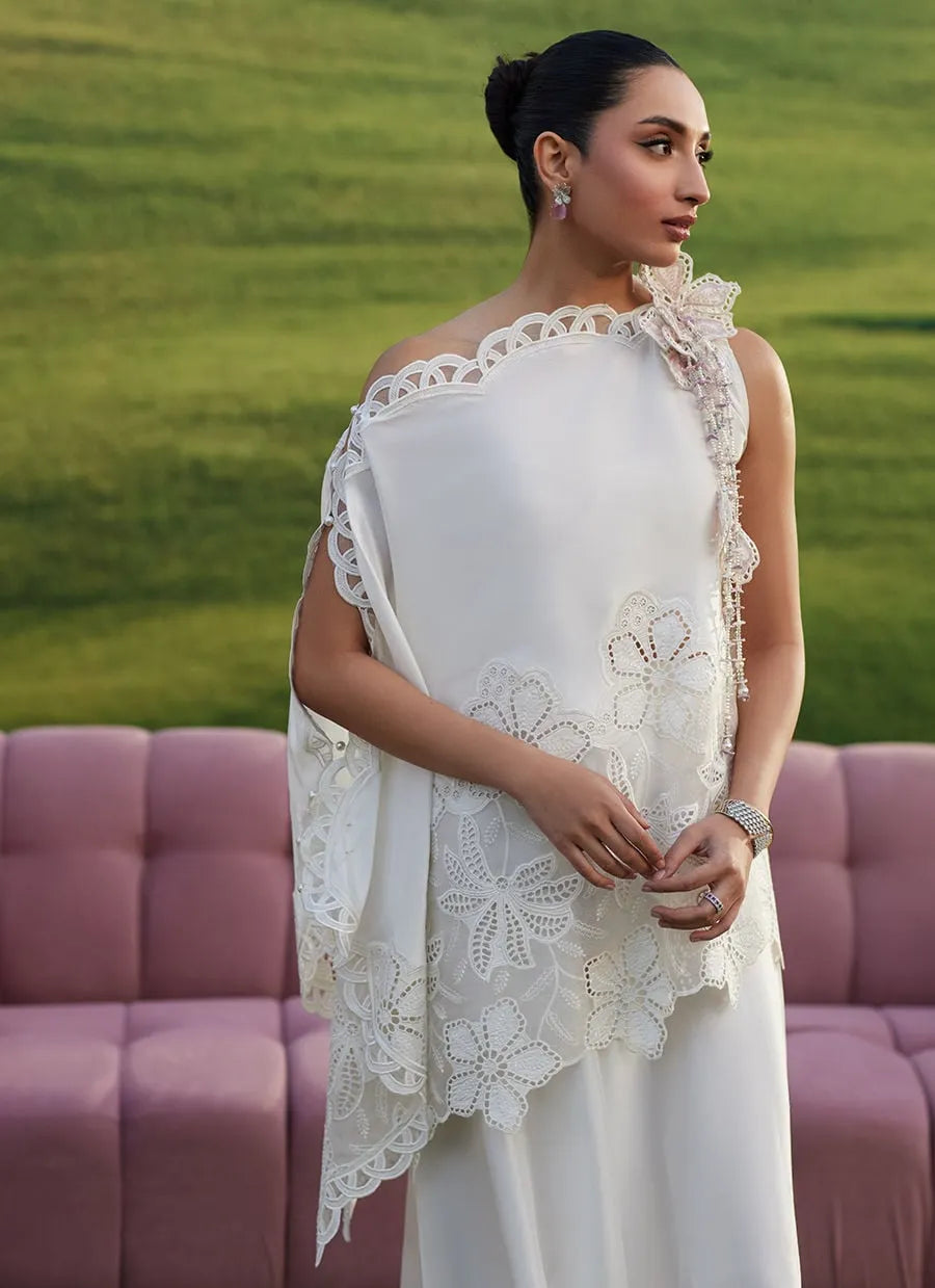 Woman in a white dress with lace details standing in front of a pink sofa and green grass.