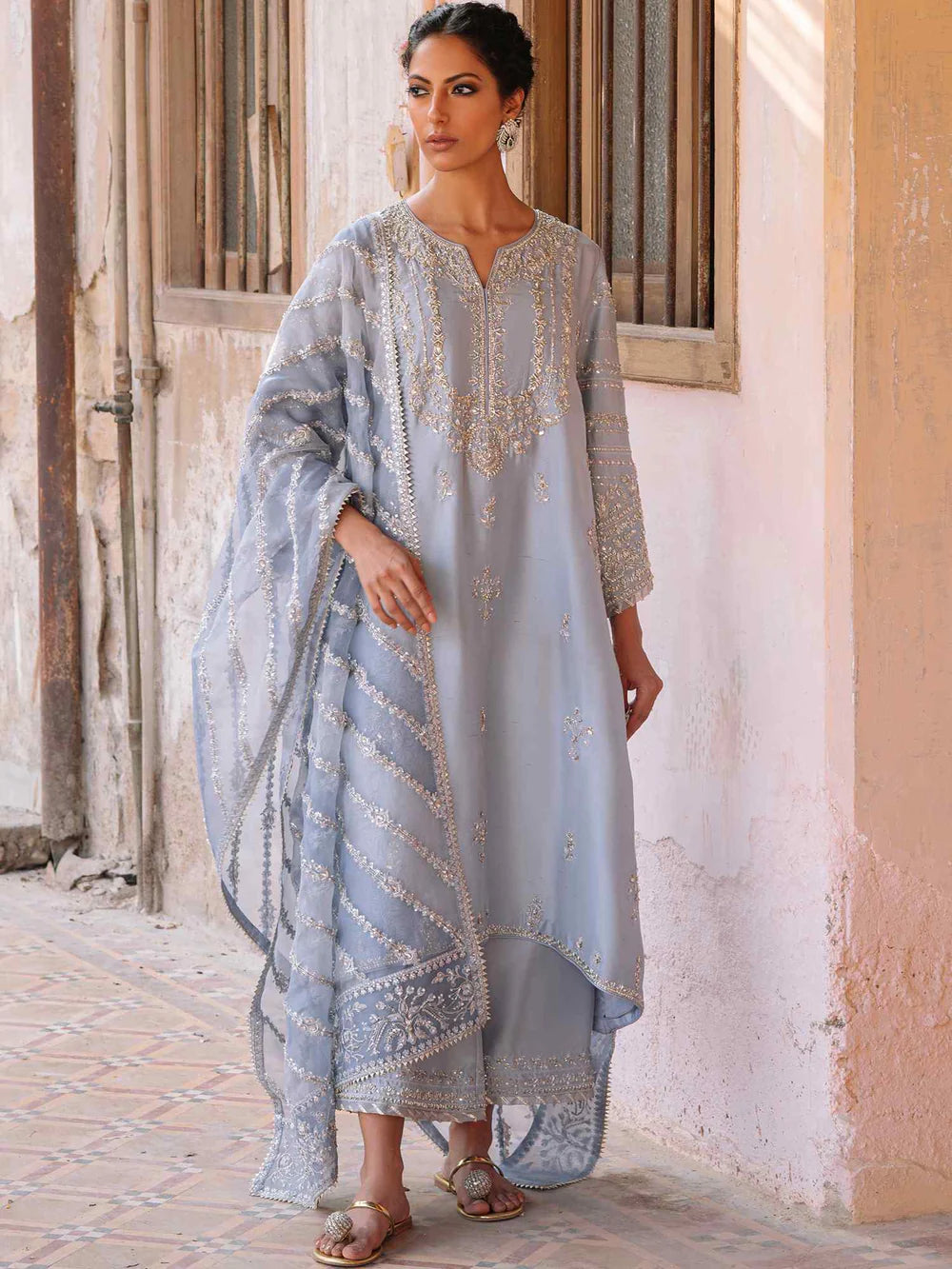 Woman wearing a light blue embroidered traditional outfit standing in front of a textured wall.