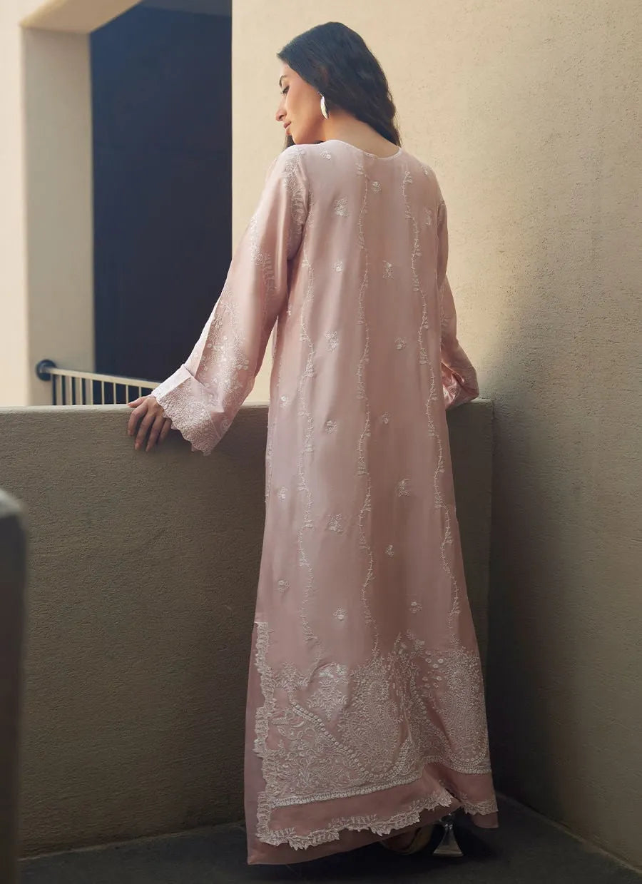 Woman in a light pink embroidered dress standing against a neutral wall.