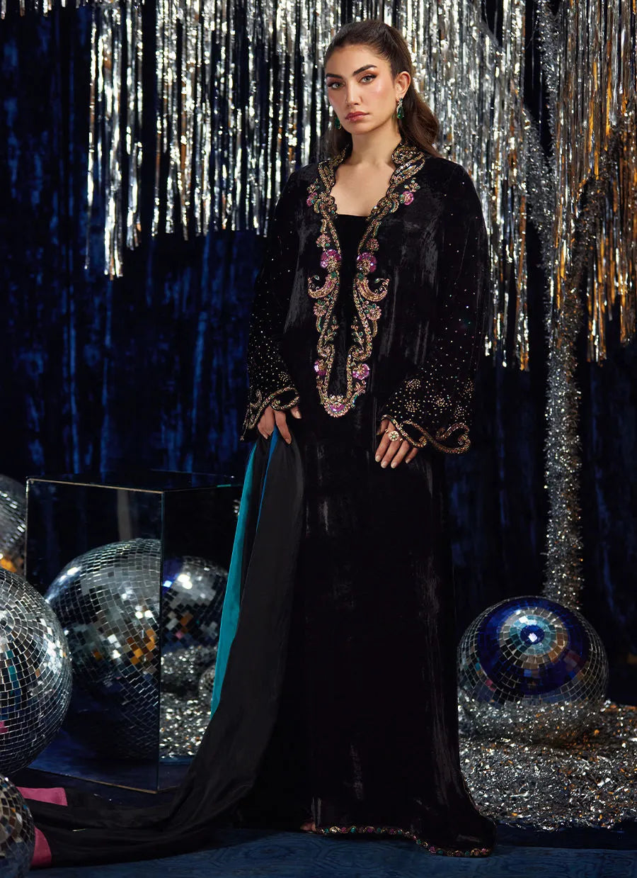 Woman in a black embroidered dress standing against a festive, glittery background.