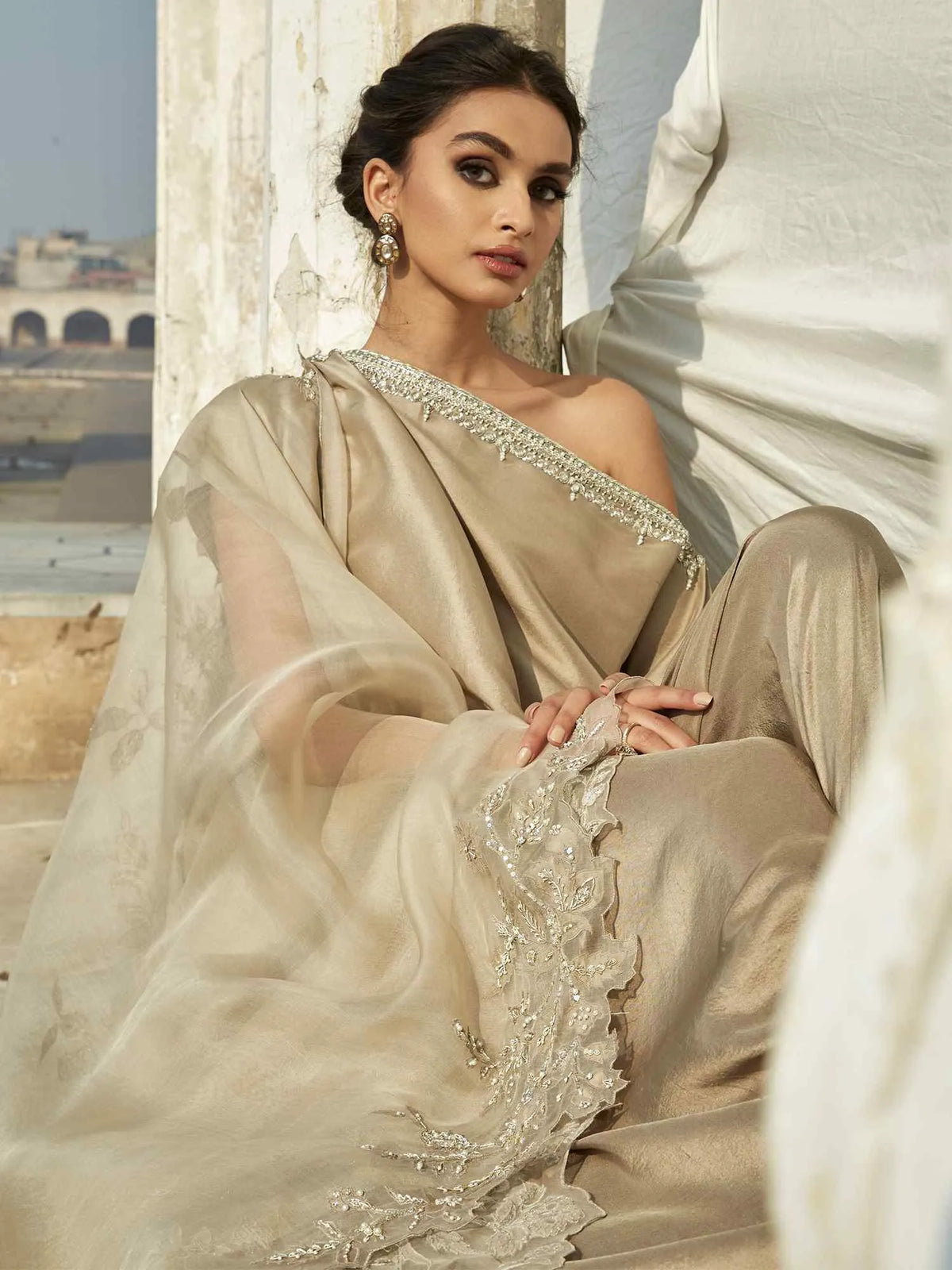 Woman in a beige saree with intricate designs, sitting outdoors.