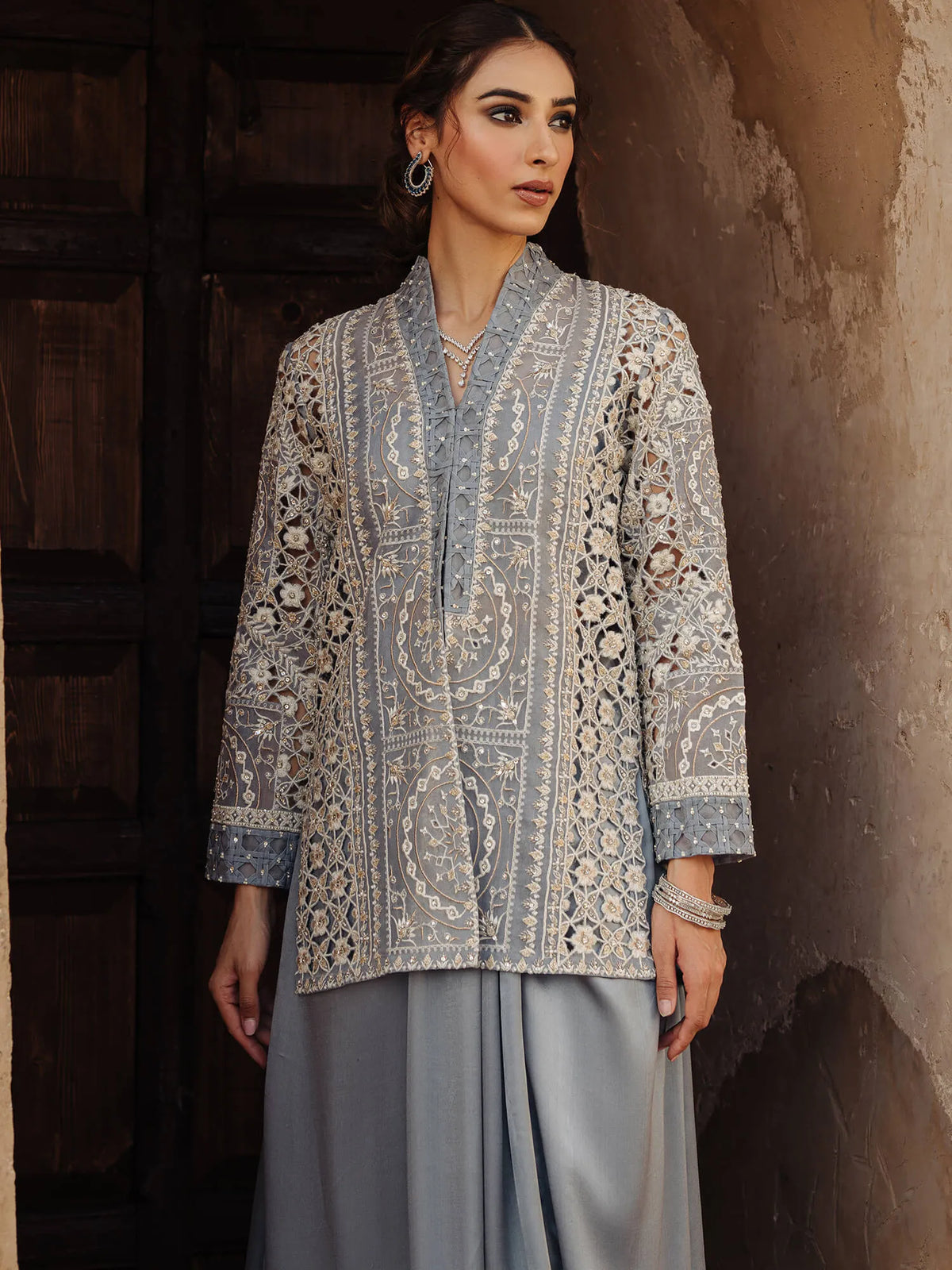 Woman wearing a patterned blouse and light gray pants against a textured wall.