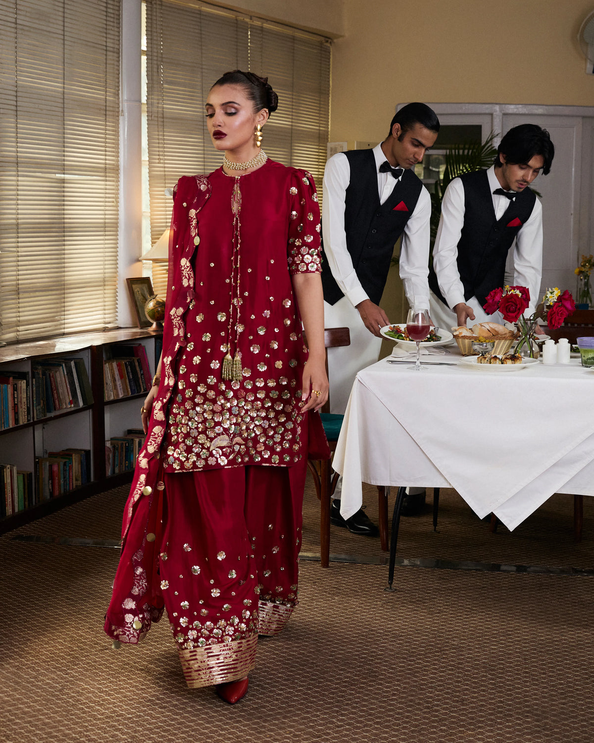 Woman in a red traditional outfit walking past a table with waiters setting it up.