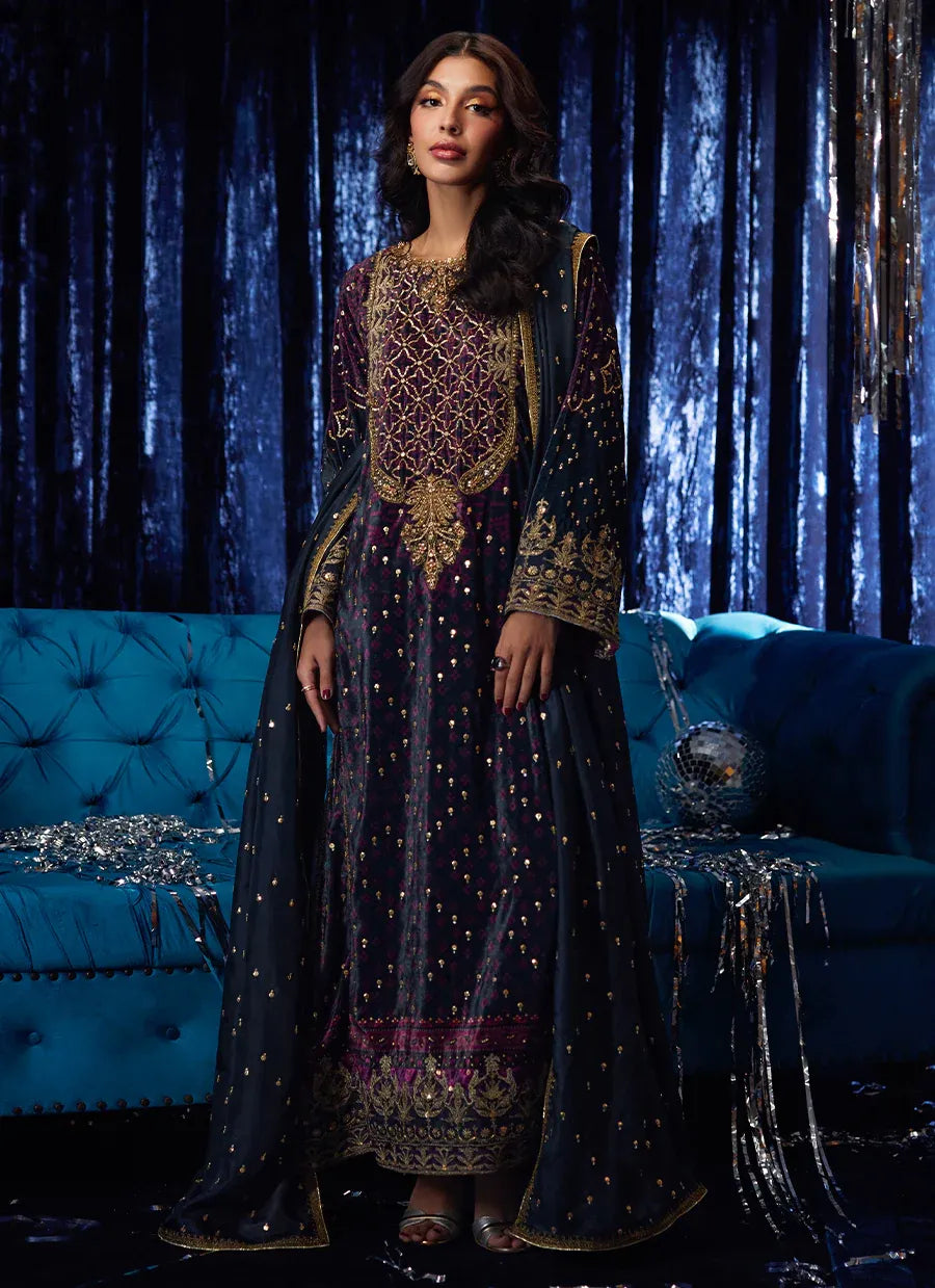 Woman in an ornate dark blue and gold dress standing in front of a blue couch with decorative elements.