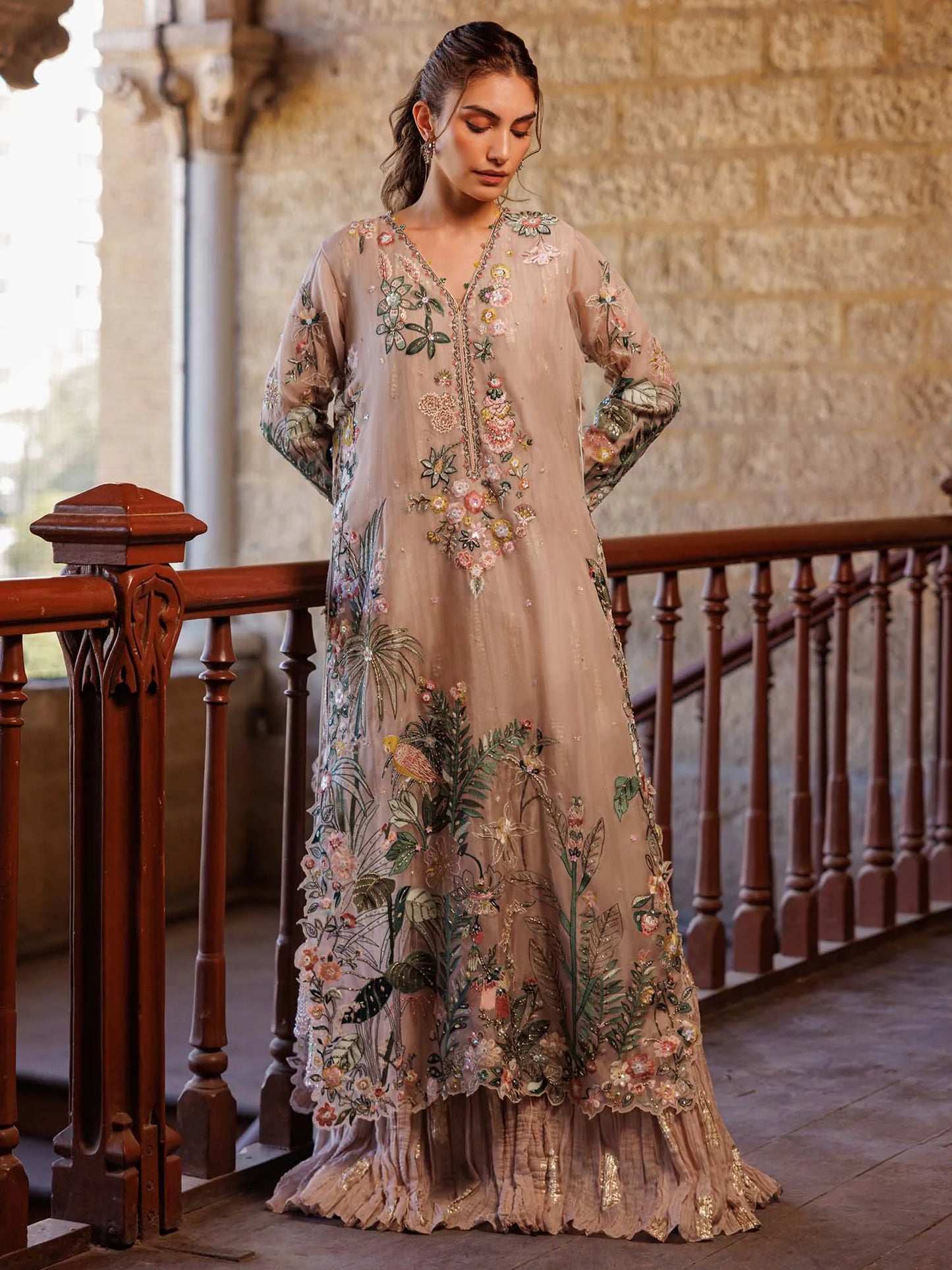 Woman wearing a floral embroidered dress in an indoor setting with stone walls and wooden railings.