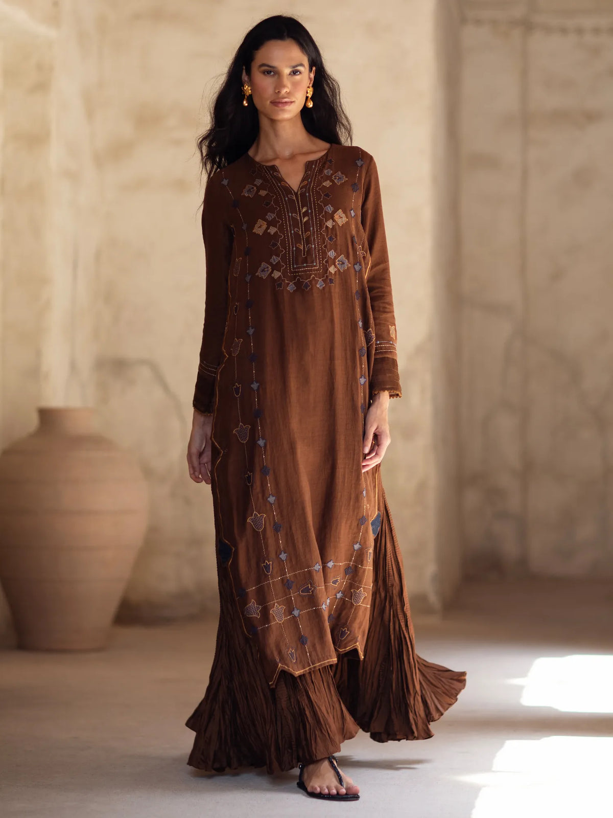 Woman wearing a long brown dress with intricate patterns in a warm-toned room.