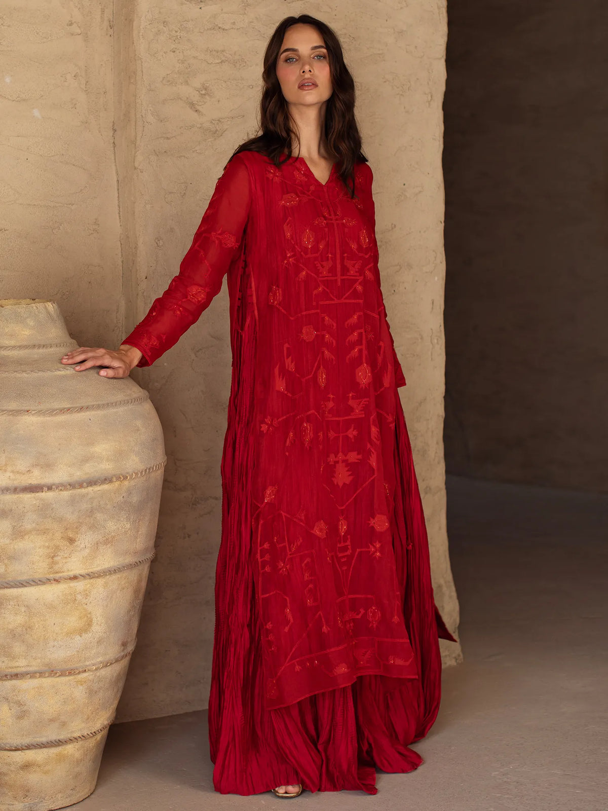 Woman wearing a red dress with intricate patterns standing next to a large stone container.