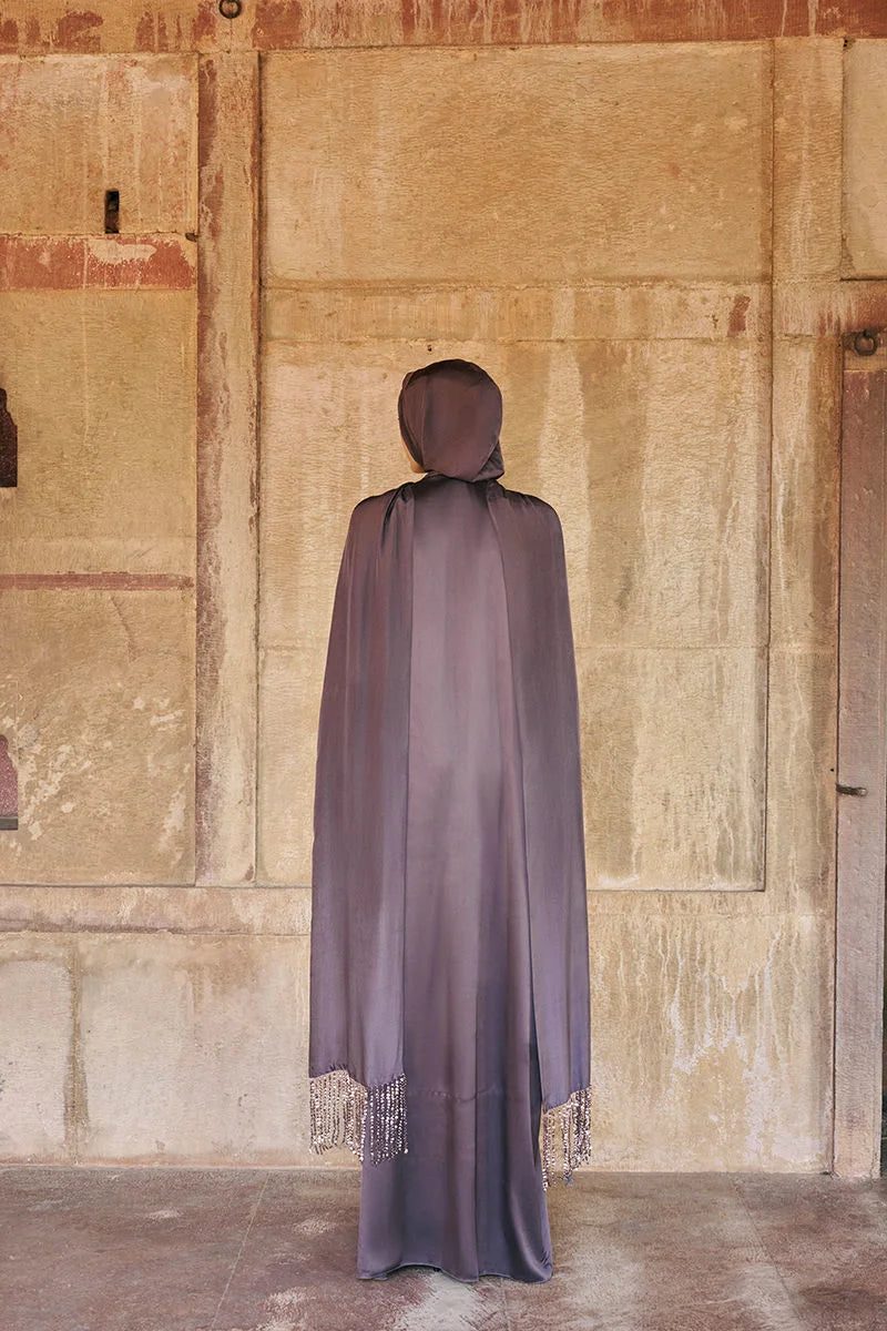 Person wearing a long, dark purple hooded garment standing against a textured beige wall.