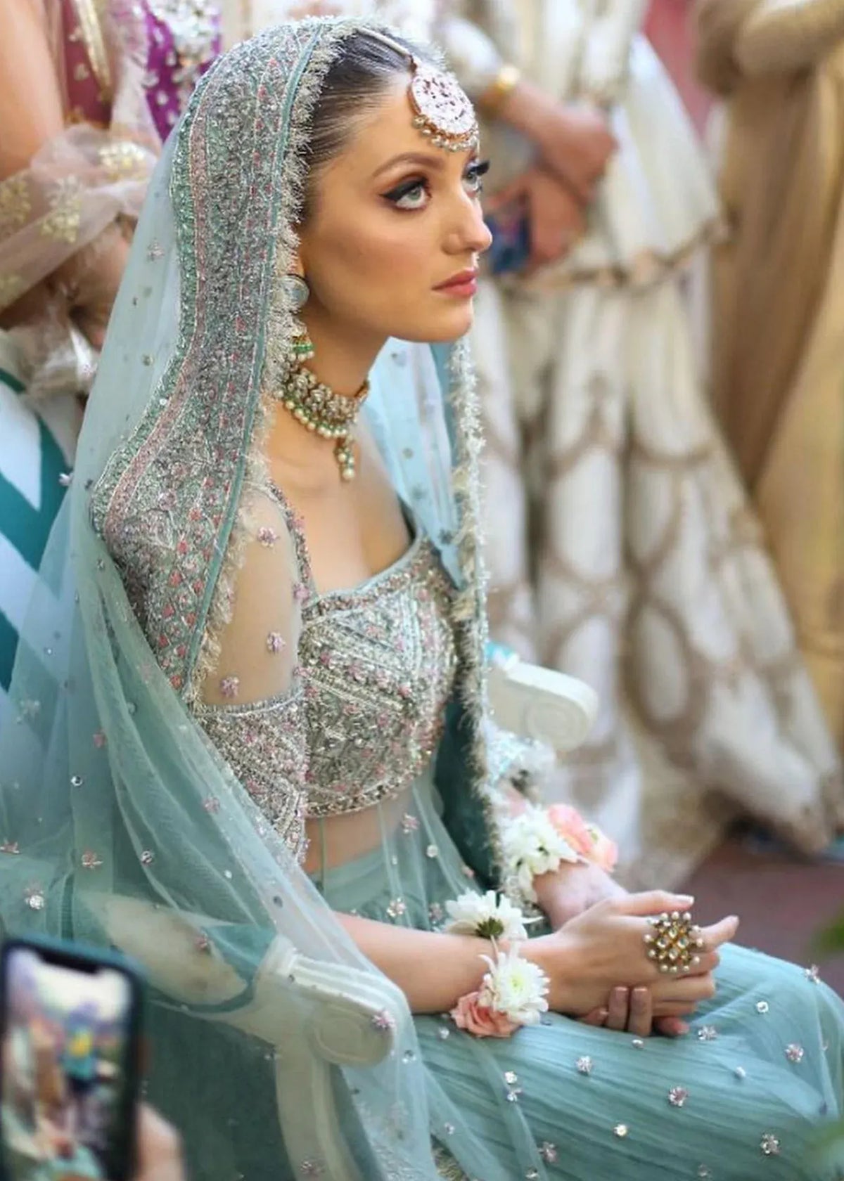 Woman in traditional attire with jewelry and a floral arrangement, surrounded by blurred people.