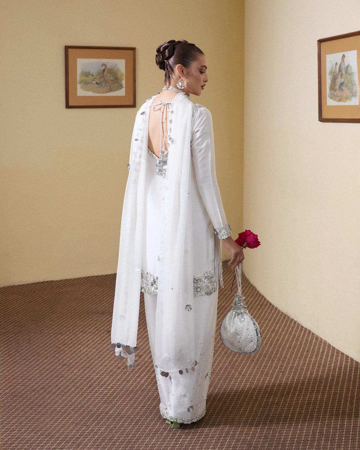Woman in a white traditional outfit with silver embroidery, holding a red flower and a matching handbag, against a beige wall.
