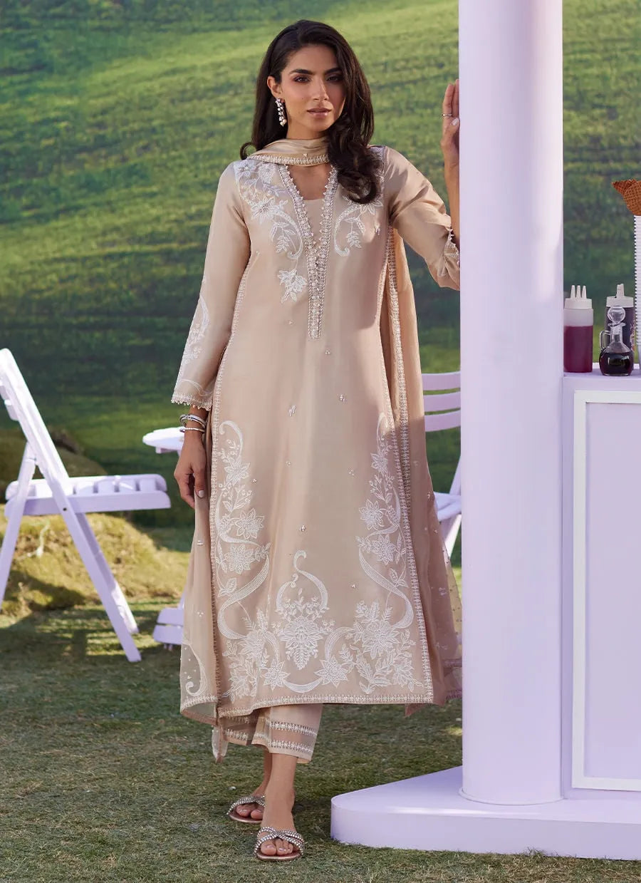 Woman in a beige embroidered outfit standing outdoors with green grass and white structures in the background.