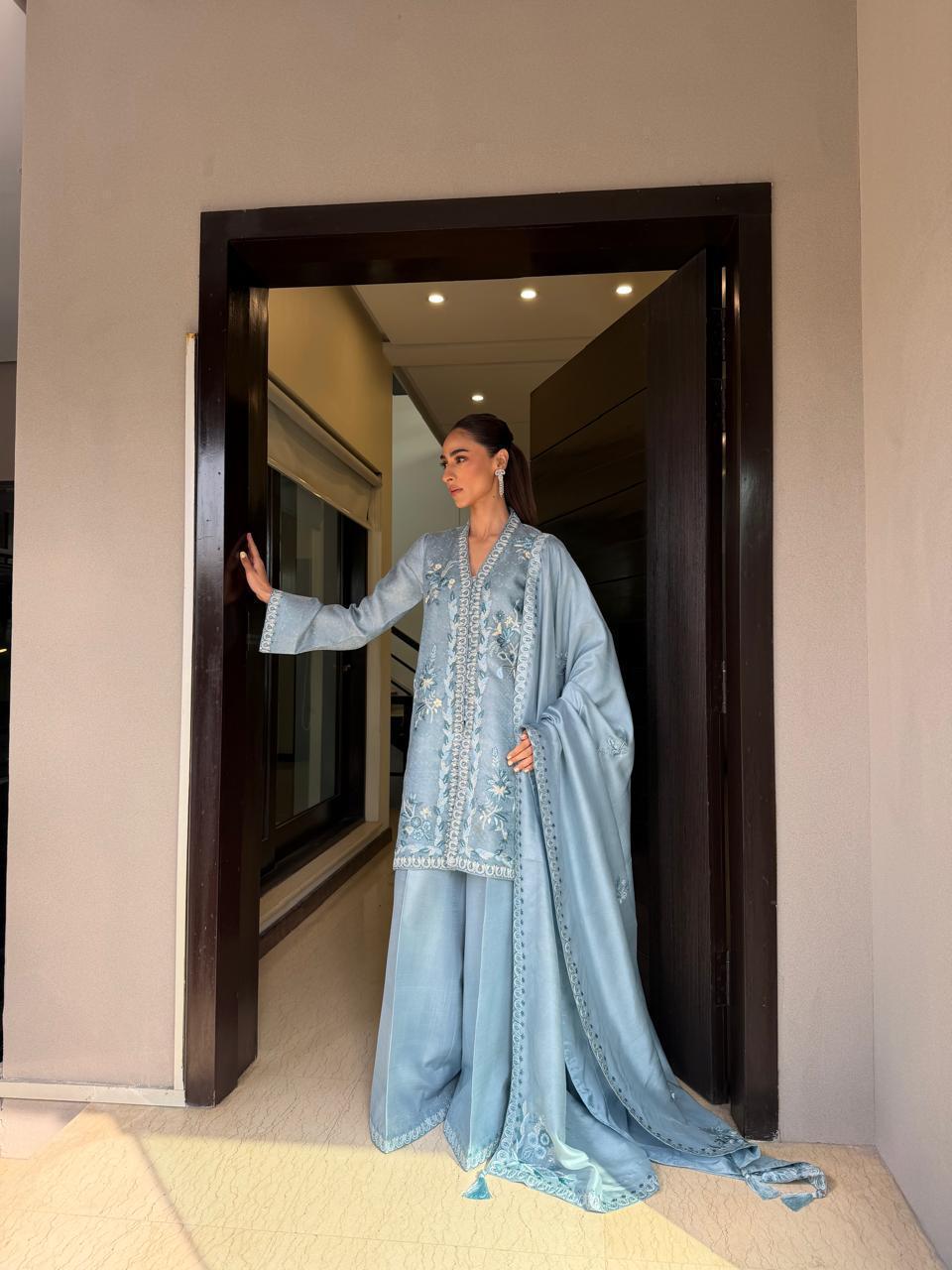 Woman in a light blue traditional outfit standing in a doorway.